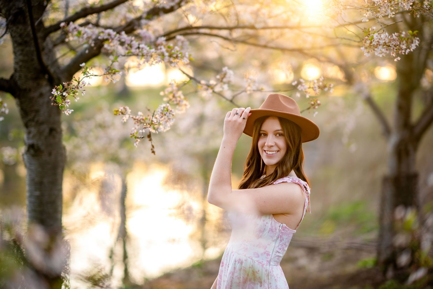 Someone in a white dress and sun hat poses beneath blooming cherry blossoms at sunset in a dreamy spring setting.