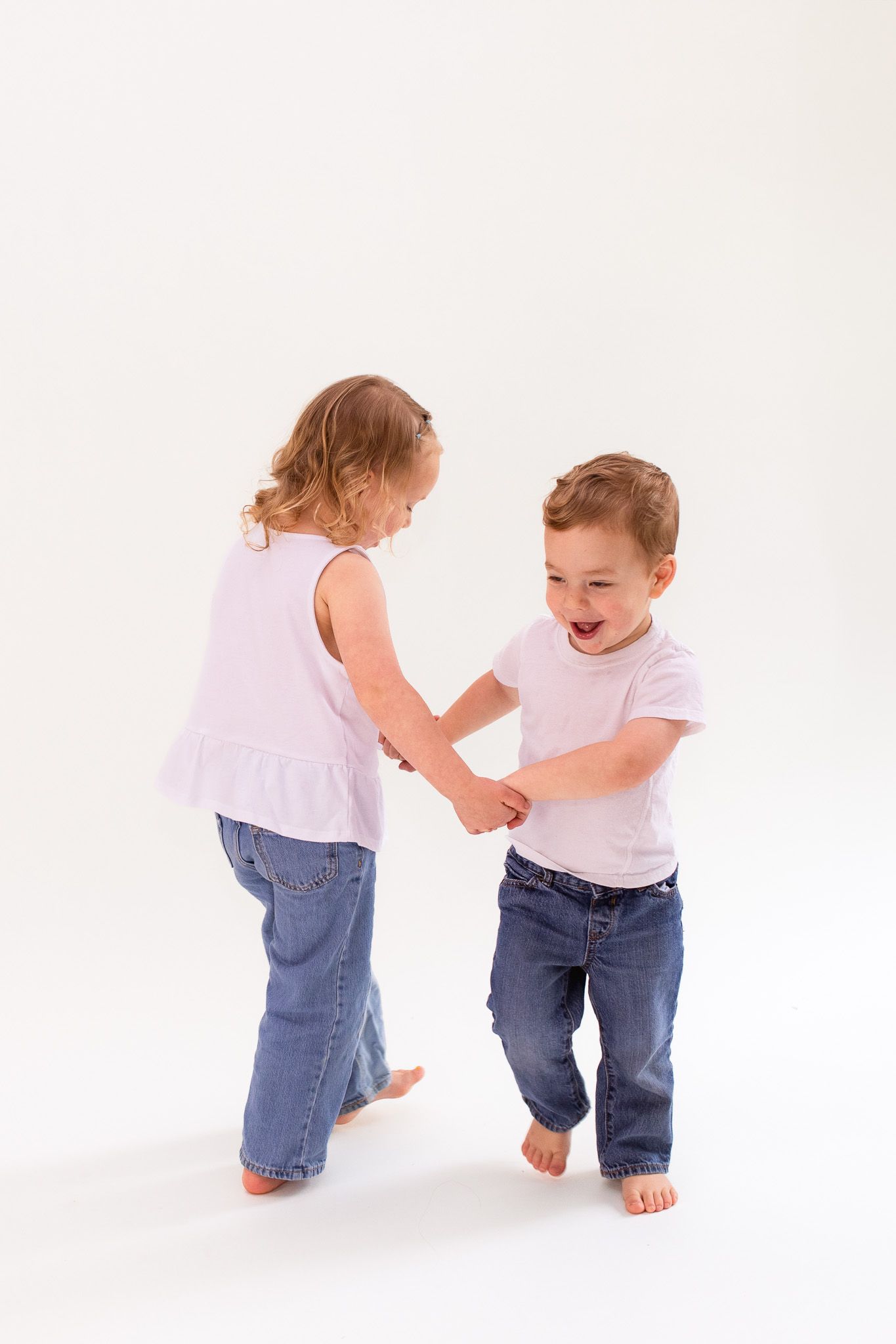 Two young children in white shirts and jeans play and laugh together on a white backdrop.