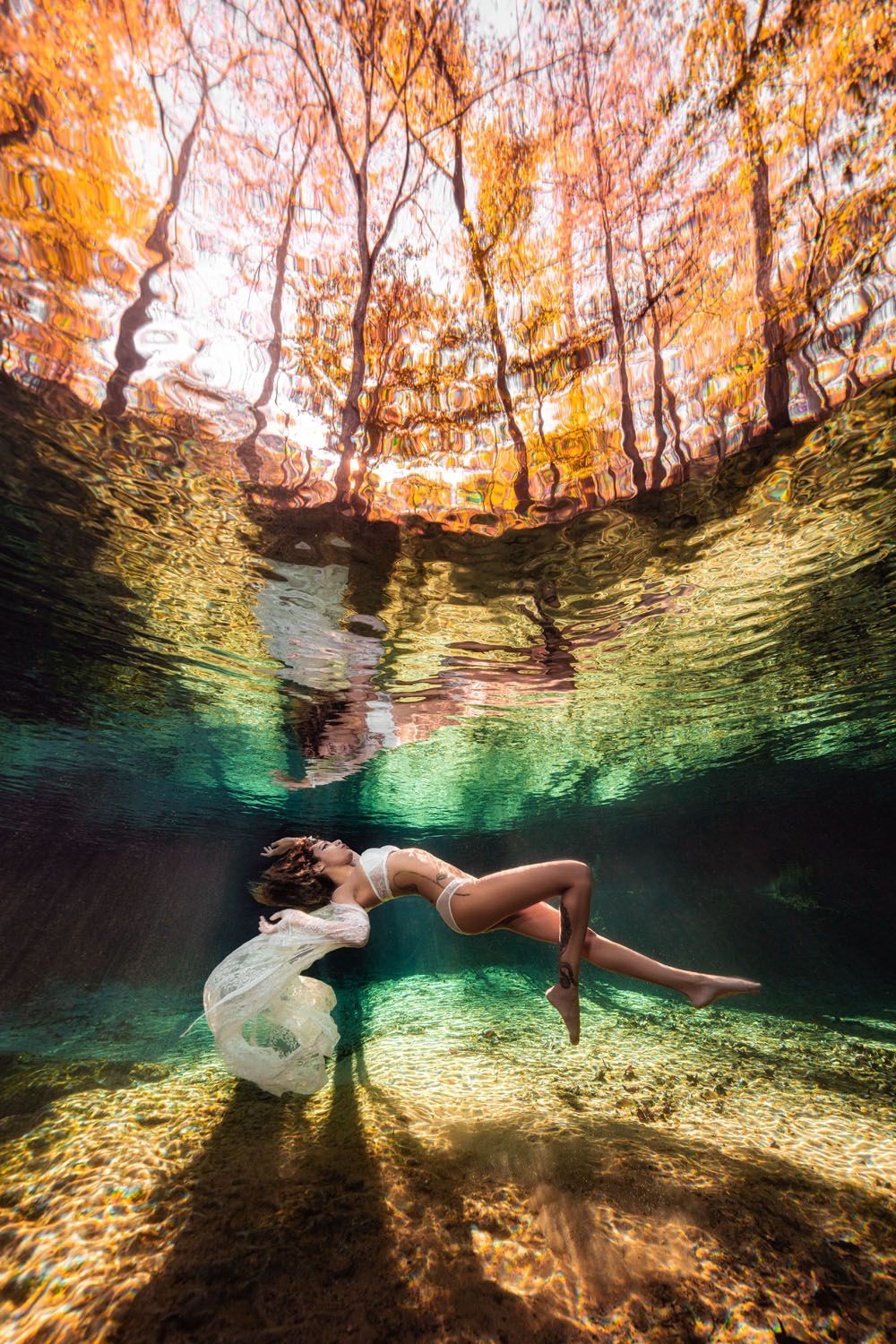 Underwater photography captures a figure in white floating ethereally in turquoise waters beneath autumn trees.