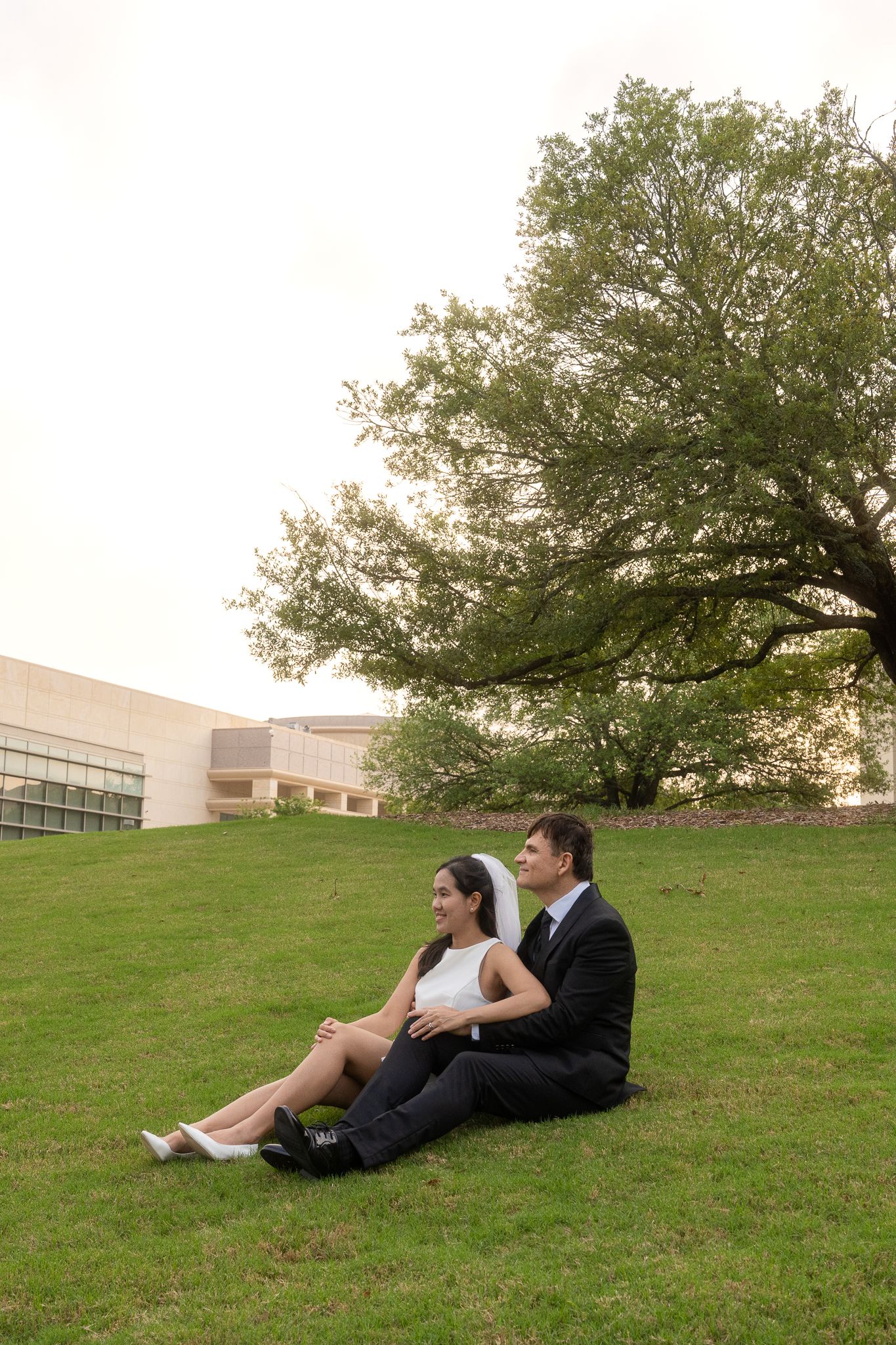 A couple sits together on a grassy hillside near a modern building during what appears to be a wedding day.