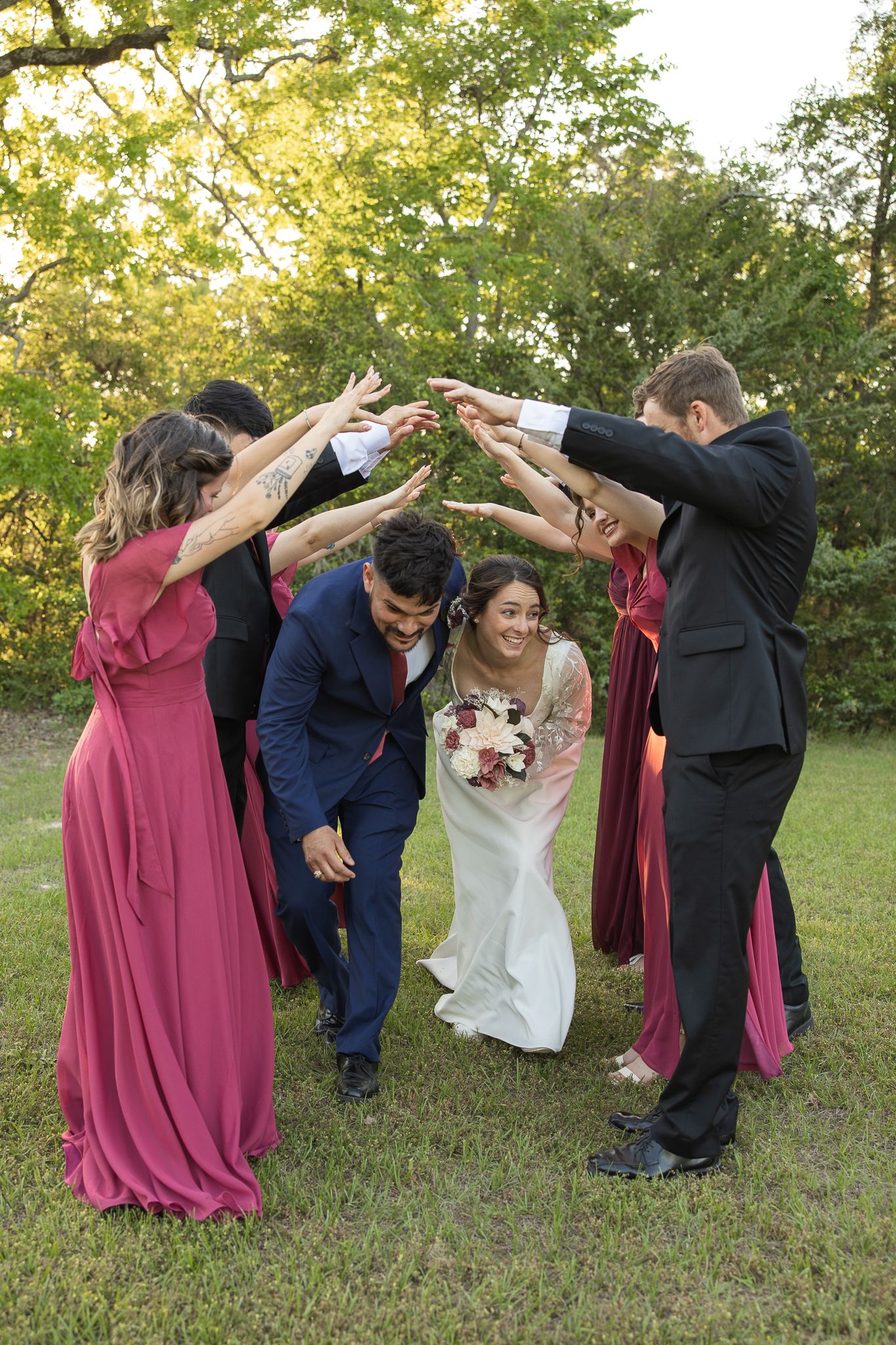 Wedding party in burgundy dresses forms an arch over couple and their dog during outdoor garden celebration.