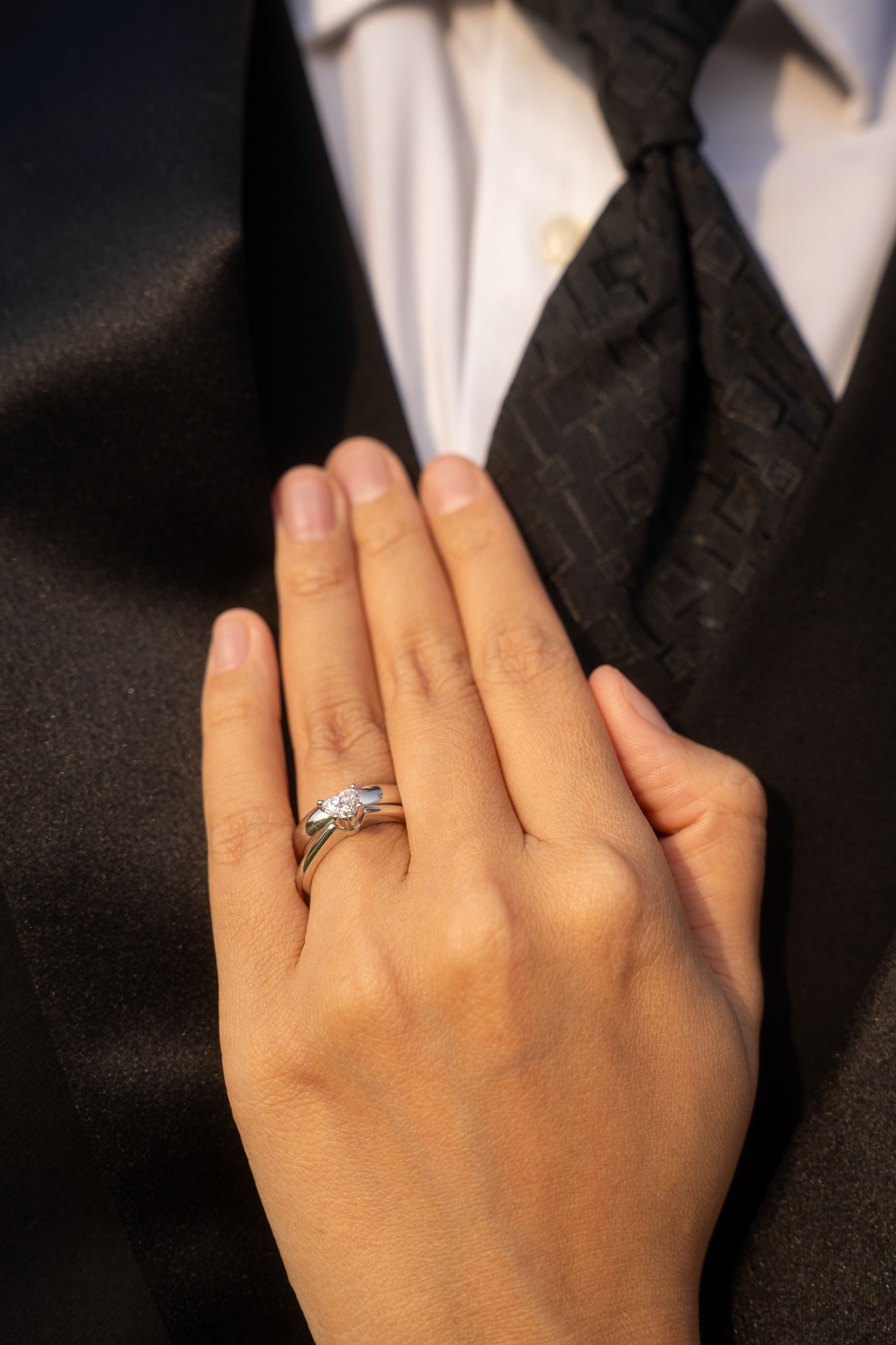 Close up of a hand wearing an elegant diamond engagement ring resting against a formal suit with black tie.
