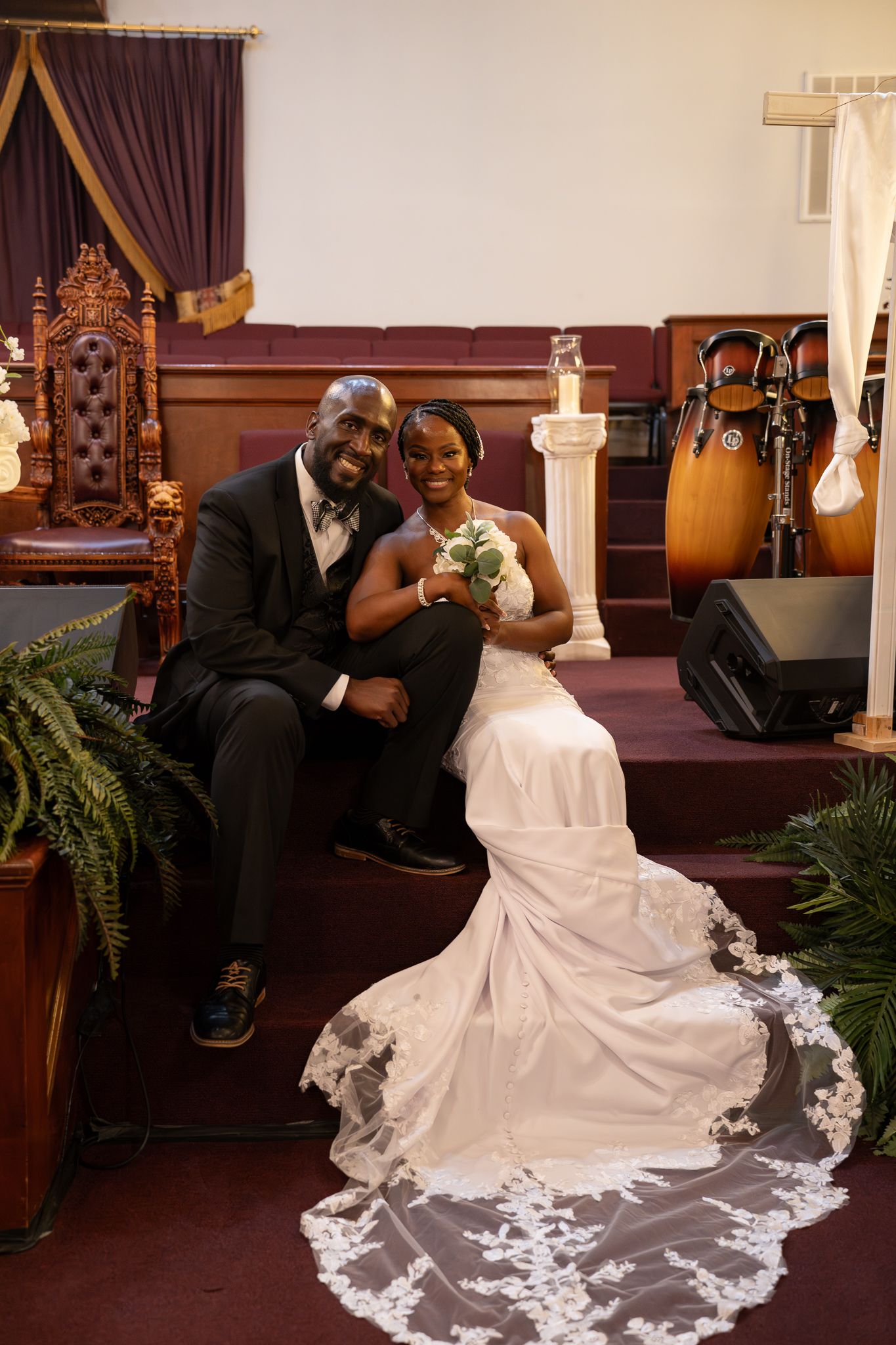 A bride in a white gown with intricate lace train poses with her groom in a luxurious church setting with dark purple curtains.