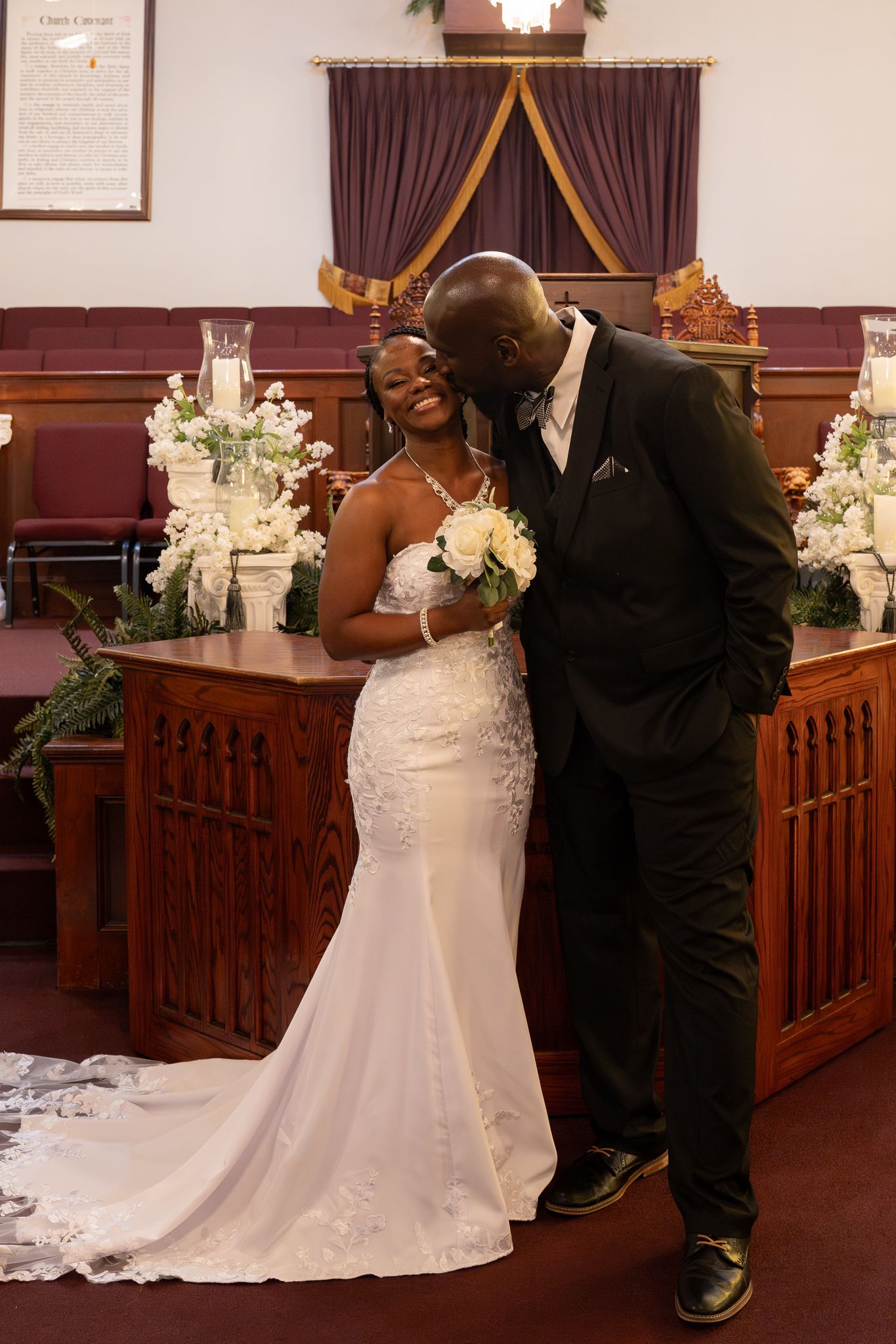 A loving wedding couple share an intimate moment at the altar surrounded by white floral arrangements in a church setting.