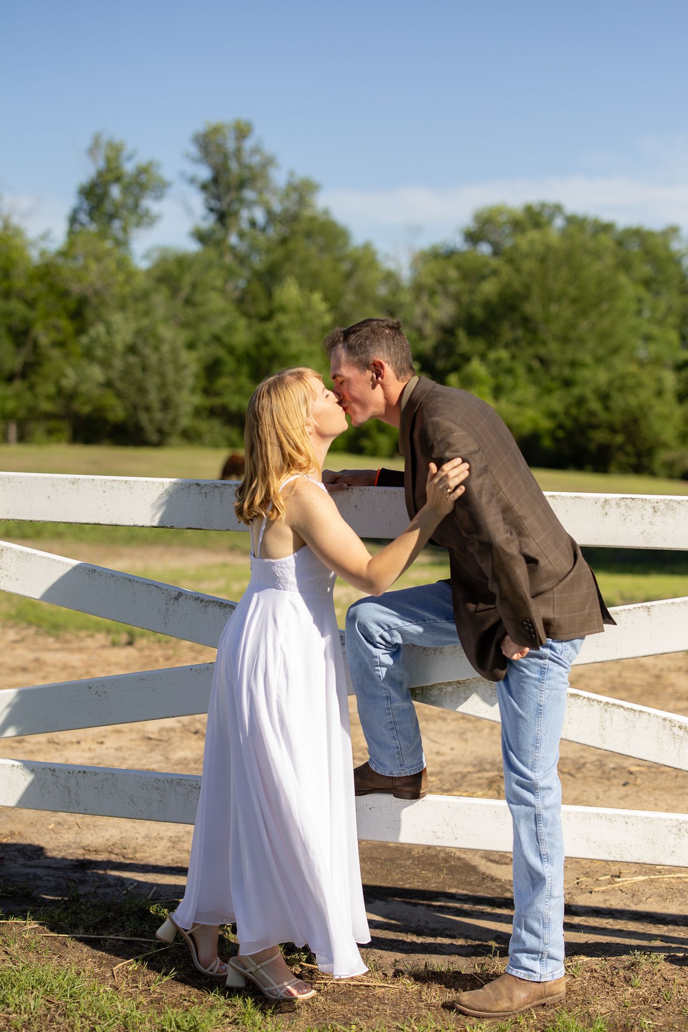 Romantic couple embraces by rustic white fence on sunny day with green trees in background.