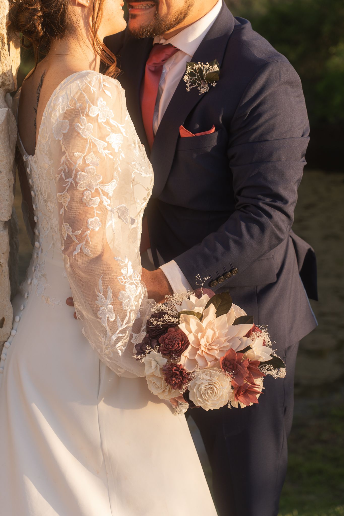 A romantic wedding detail shot showing navy suit and lace gown, with a burgundy and white floral bouquet in soft sunlight.