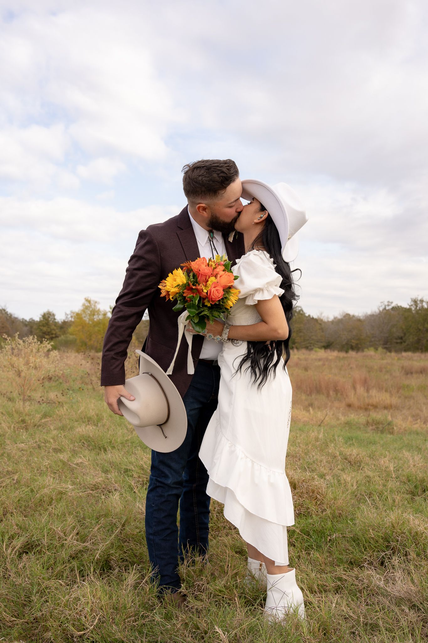 A couple shares a romantic kiss in a field while holding a vibrant autumn bouquet and cowboy hat.