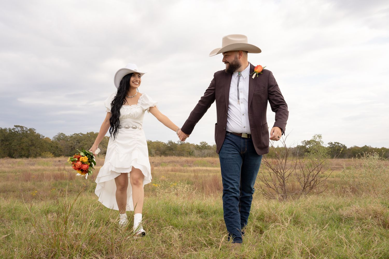A couple in western wedding attire walks hand in hand through a rustic field with autumn flowers.