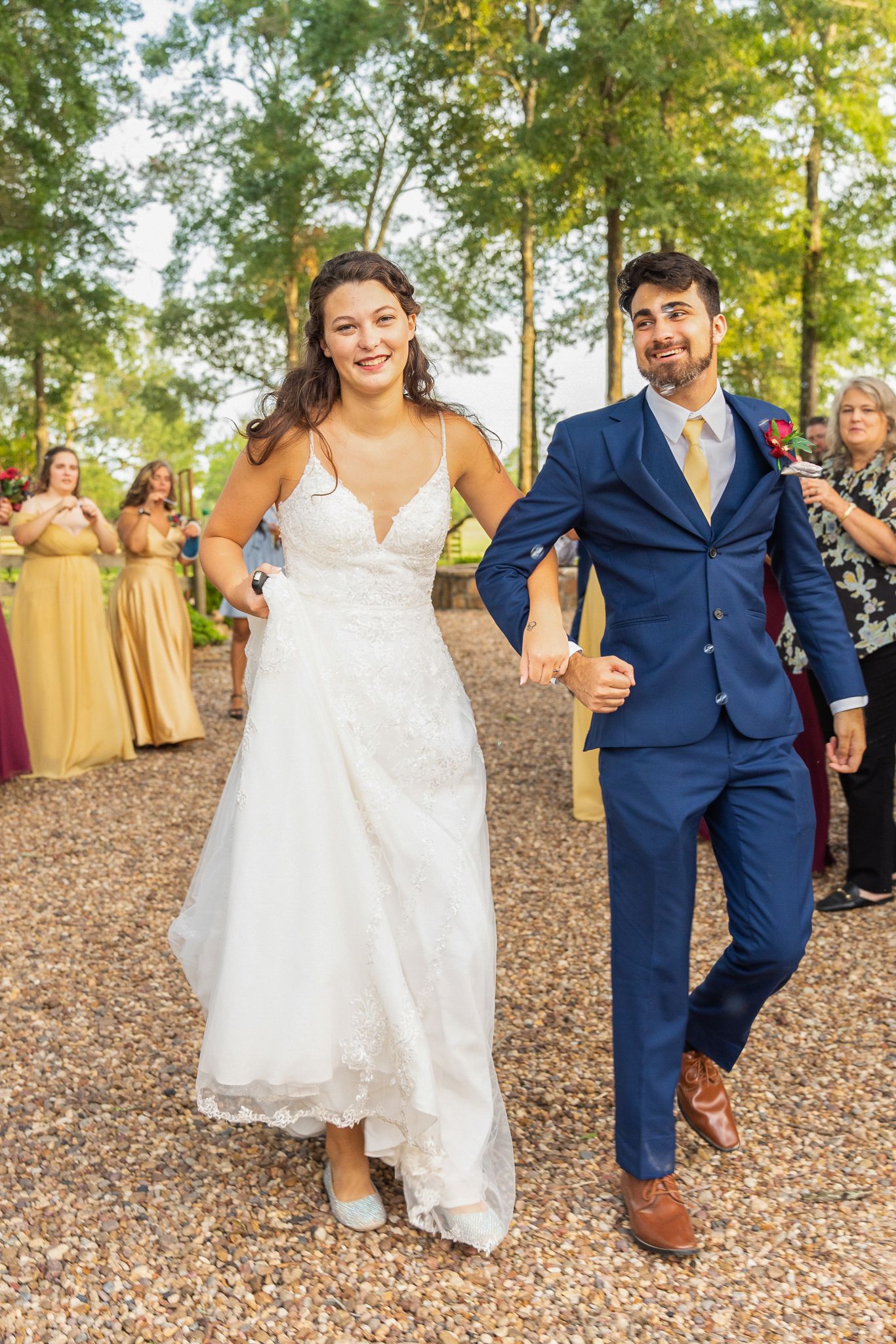 Newlyweds walk together on a gravel path with bridesmaids in gold dresses visible in the background among trees.