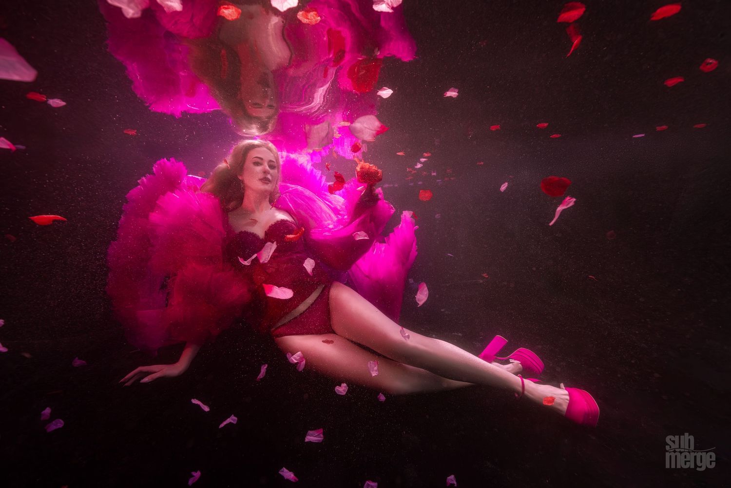 Figure in pink flowing dress poses gracefully underwater surrounded by floating petals in magenta-lit scene.