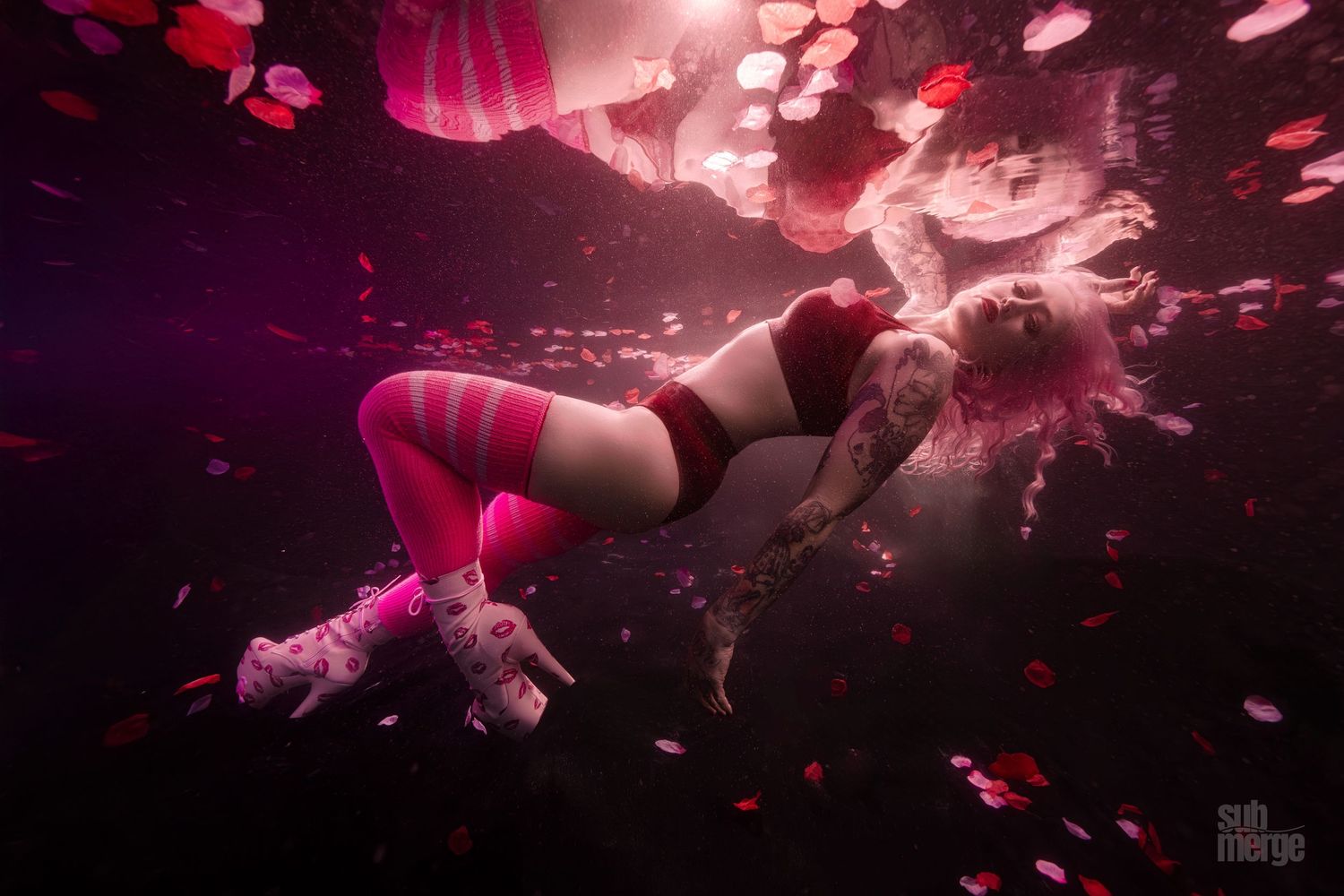 Underwater figure drifts in pink-lit water surrounded by scattered petals creating a dreamy, ethereal atmosphere.
