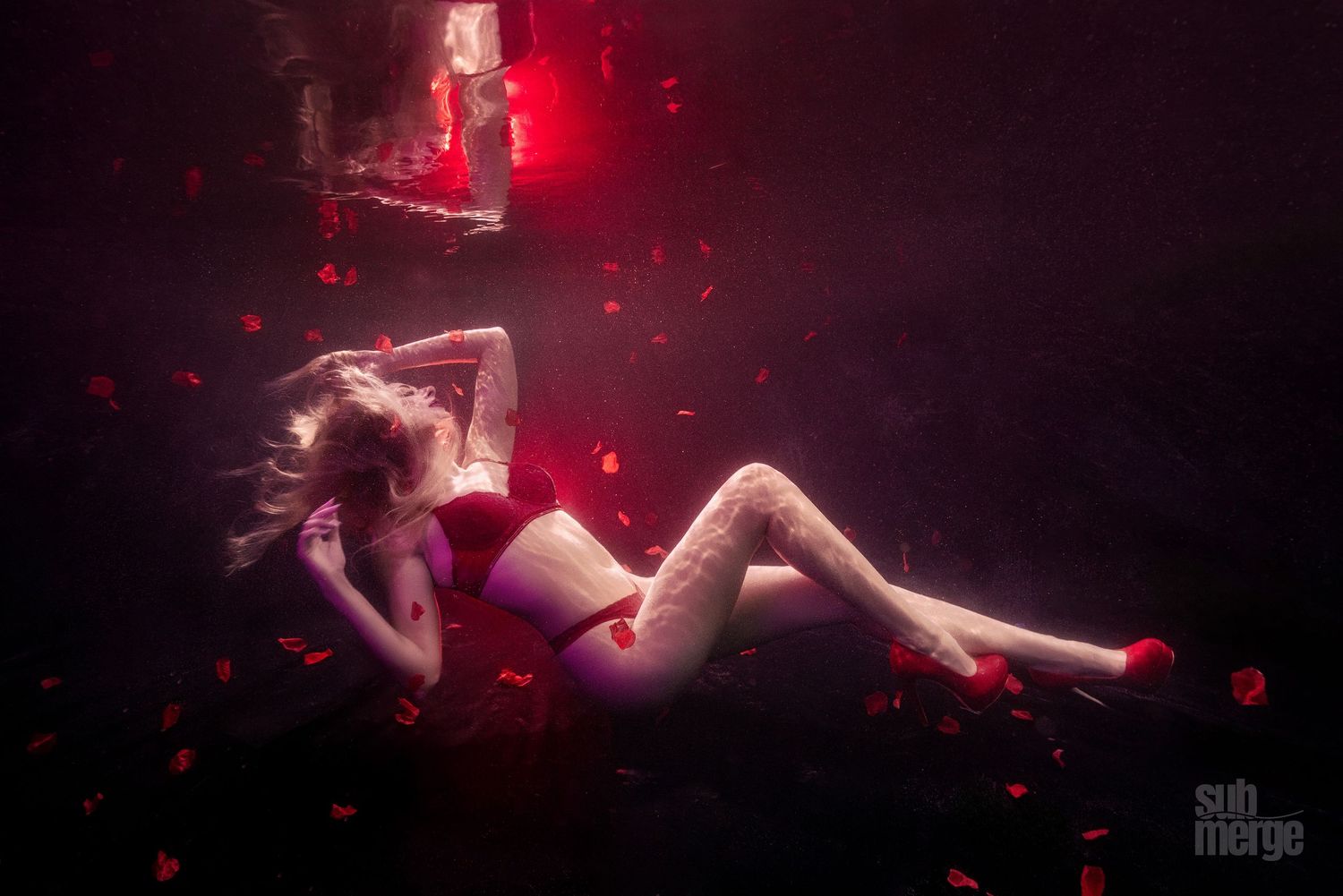 An artistic underwater portrait with floating rose petals and dramatic red lighting creates a dreamy atmosphere.