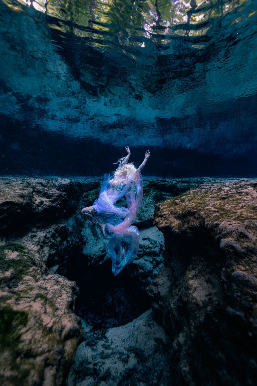 Ethereal underwater scene in turquoise cenote with a floating figure draped in flowing purple fabric against dark rocky walls.