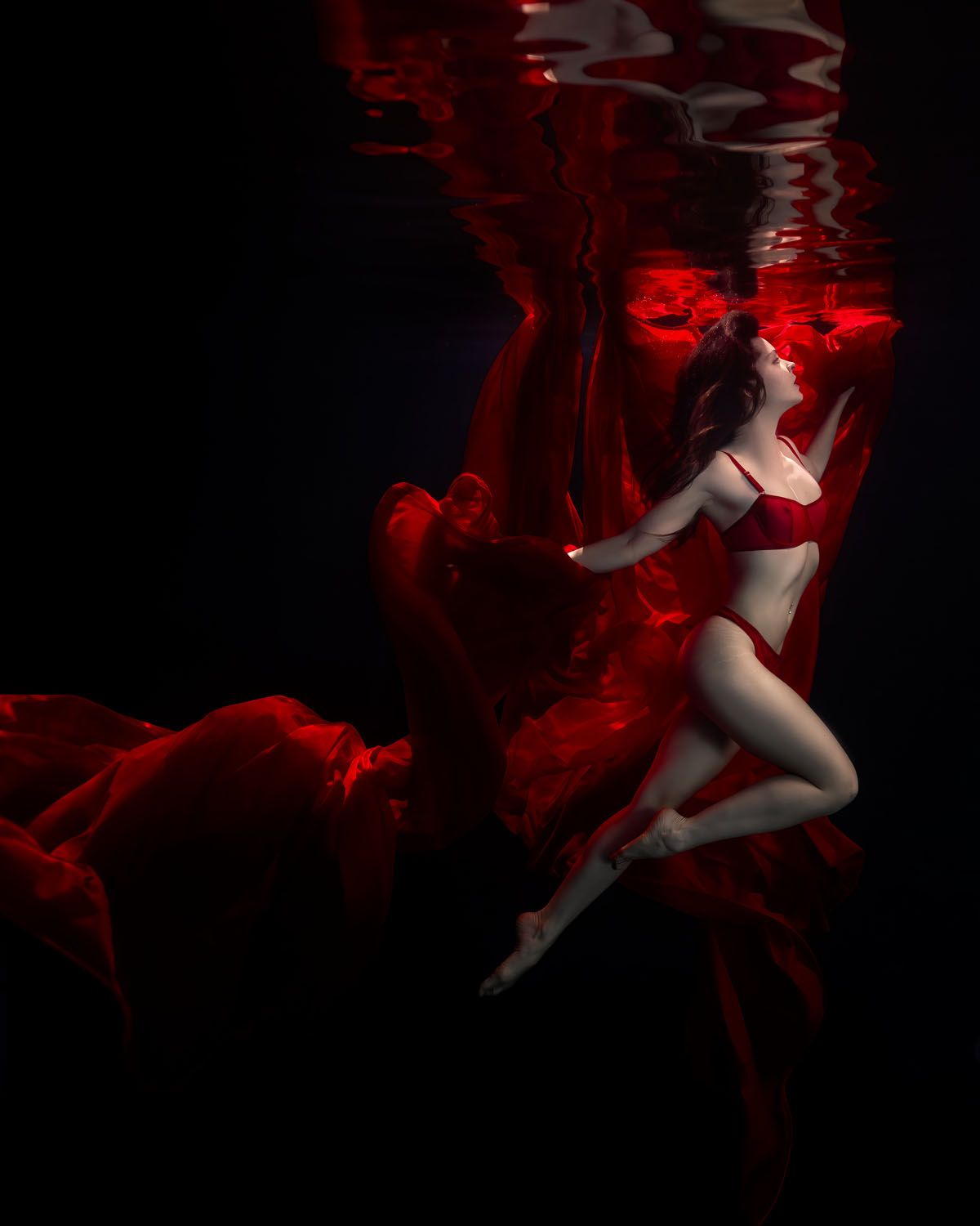 Figure in white swimwear suspended underwater with flowing red fabric creating dramatic ethereal effects in dark setting.