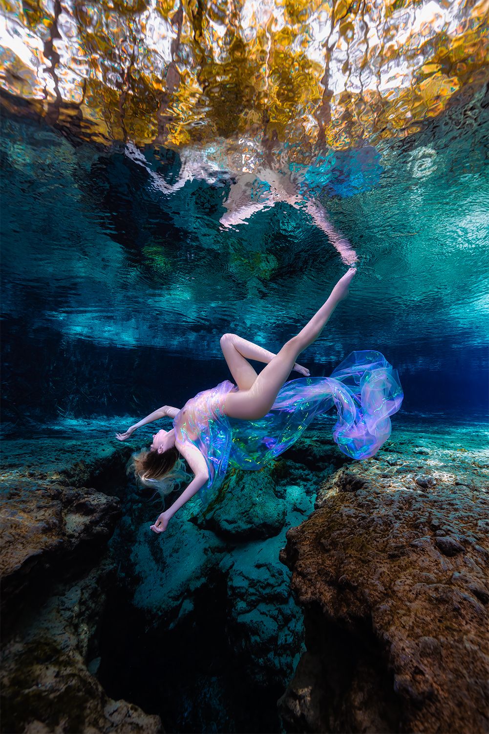 Artistic underwater photograph of a figure gracefully floating in turquoise blue cenote waters with golden reflections above.