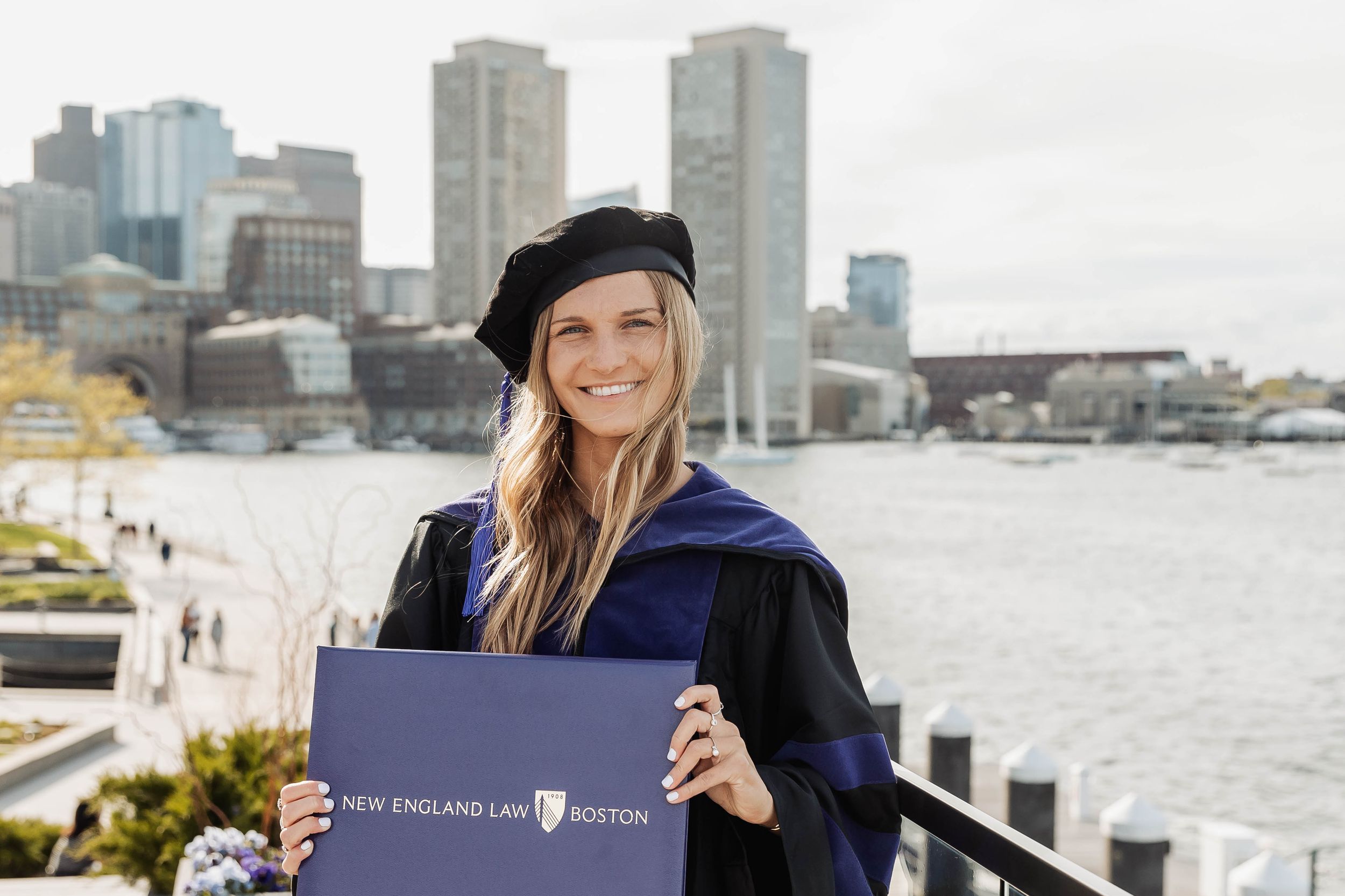 Graduate student poses with diploma on waterfront with Boston city skyline and harbor in background.