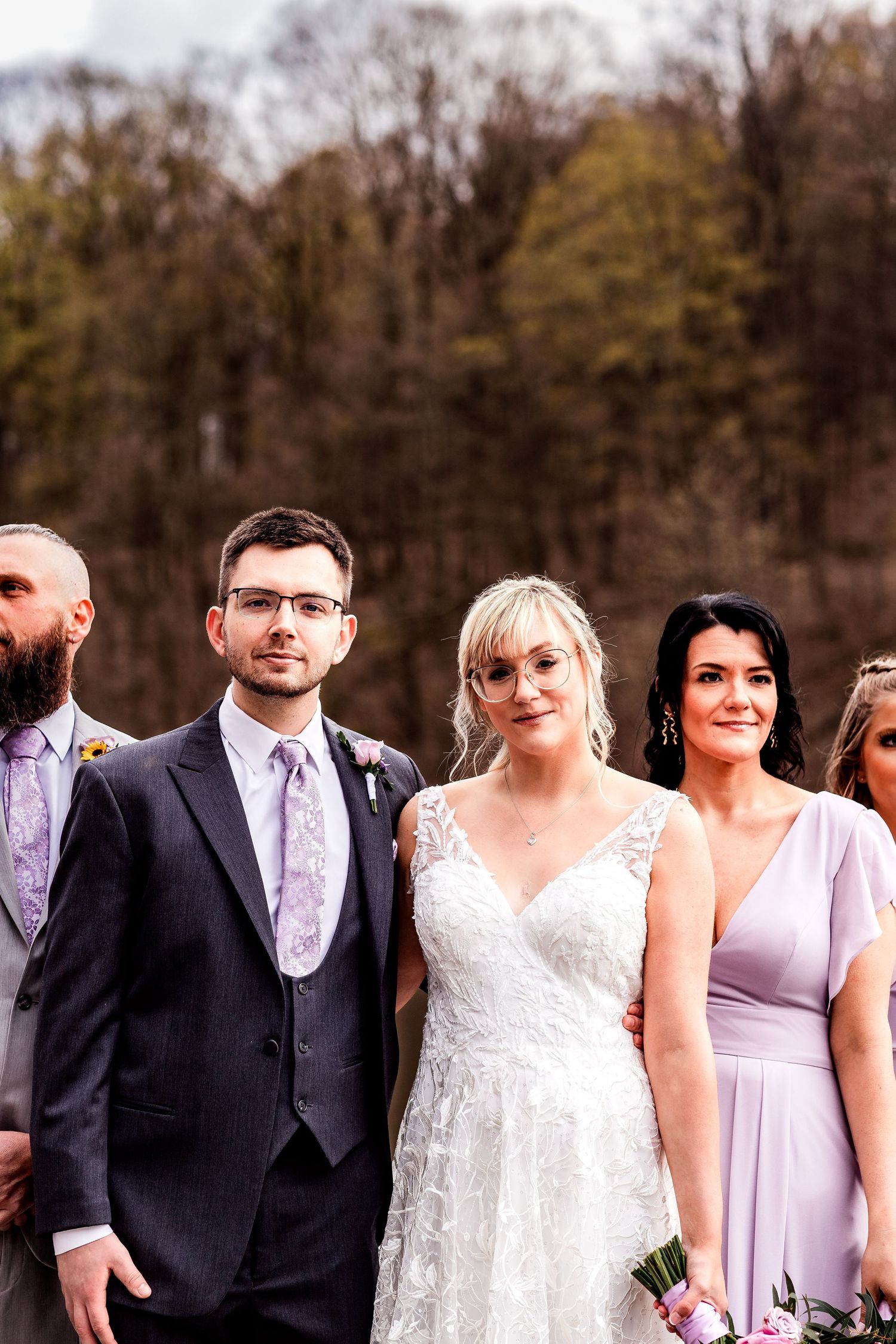 Wedding party members in formal attire gather outdoors with autumn trees in the background.