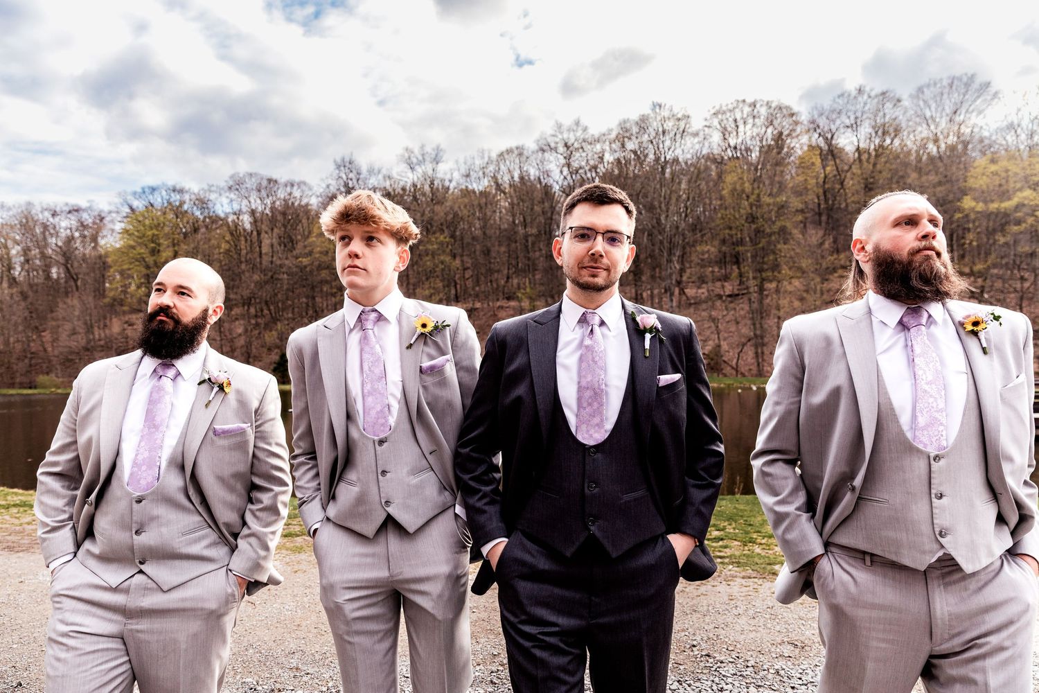 Four groomsmen in formal suits pose together near a lake with light gray suits and a navy suit.