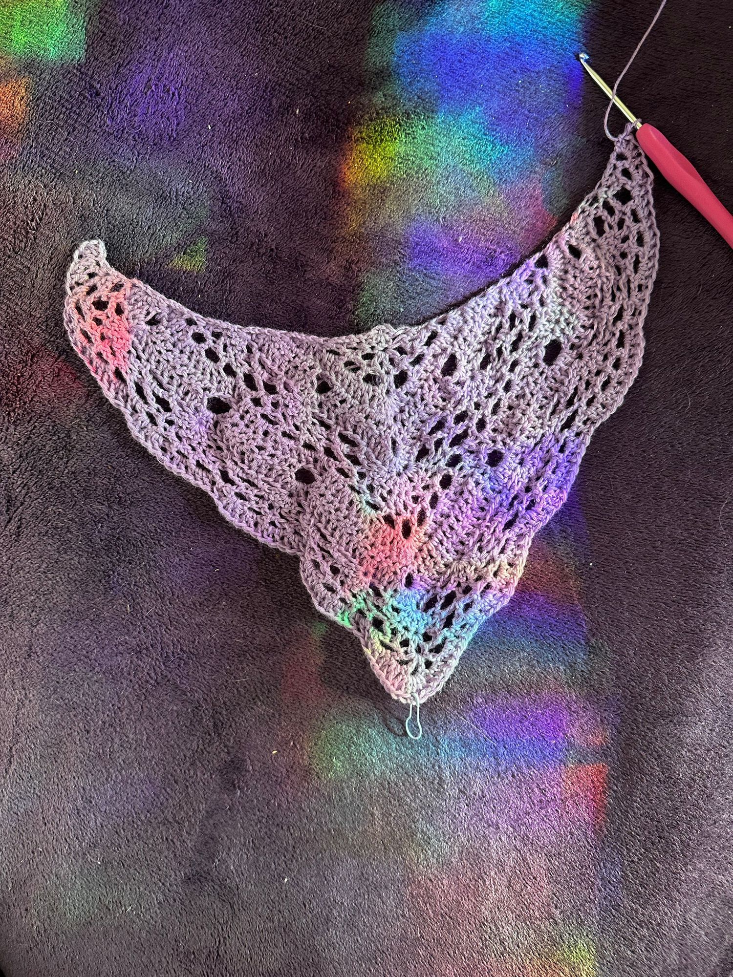 Delicate purple crochet lace shawl with intricate pattern against dark background with colorful rainbow reflections.