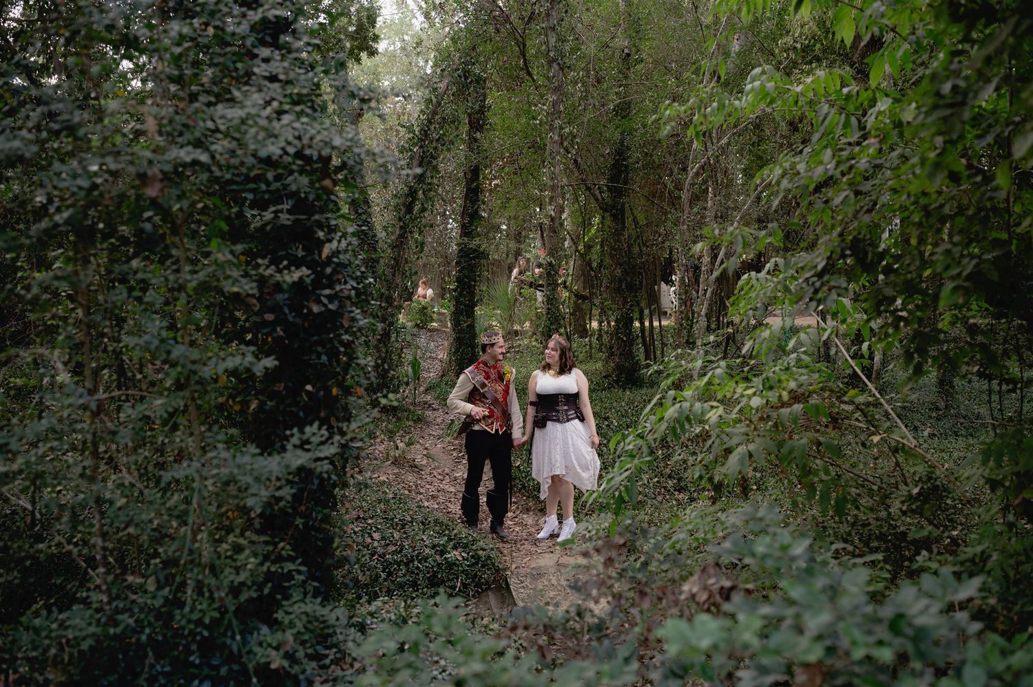 A group is seen hiking through a scenic wooded trail surrounded by lush green foliage and tall trees.