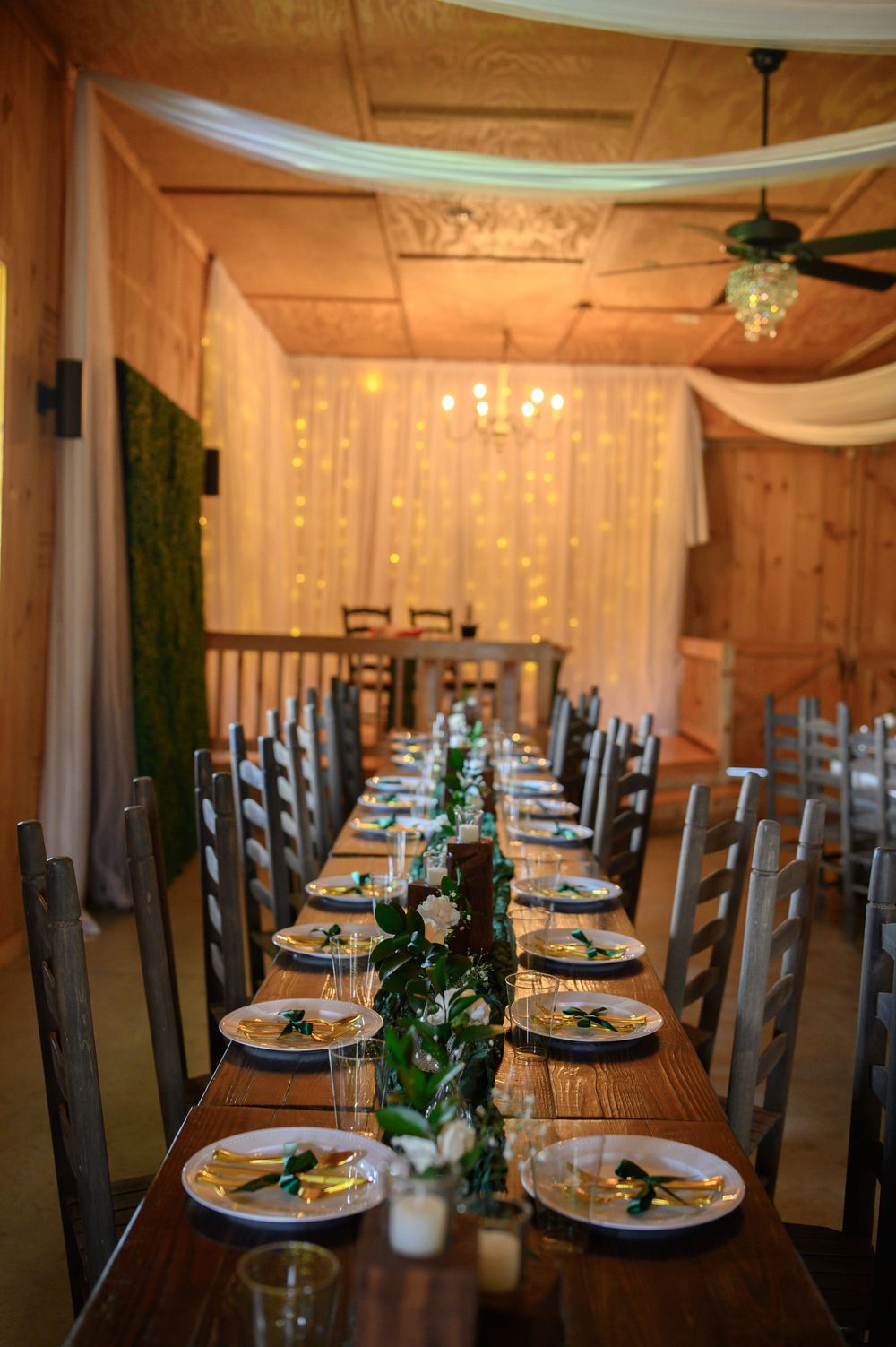 A rustic wedding reception table with wooden decor, white draping, fairy lights and elegant place settings in warm lighting.