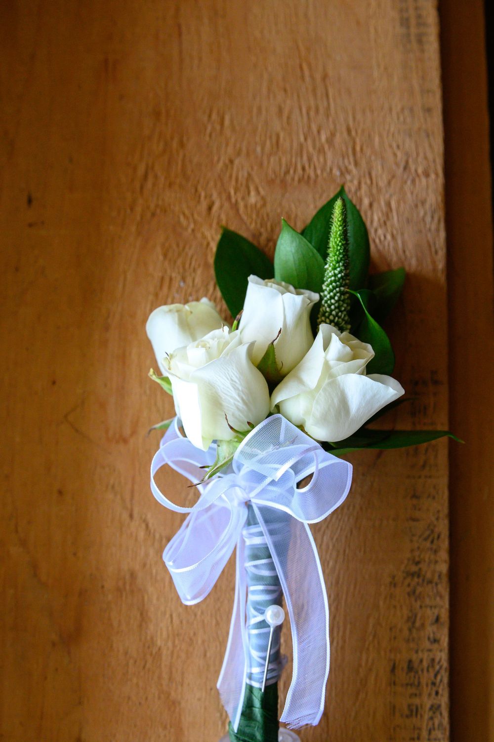 A white rose boutonniere with green leaves and a white bow displayed against a brown wooden surface.