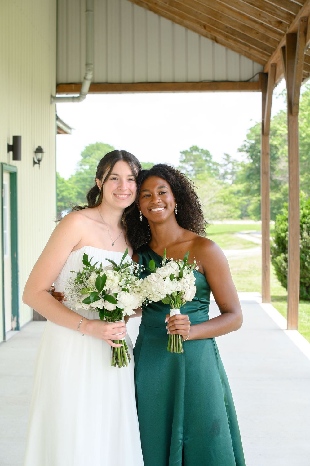 Bridesmaids in emerald green dresses pose with bride in white wedding gown carrying bouquets along covered walkway.