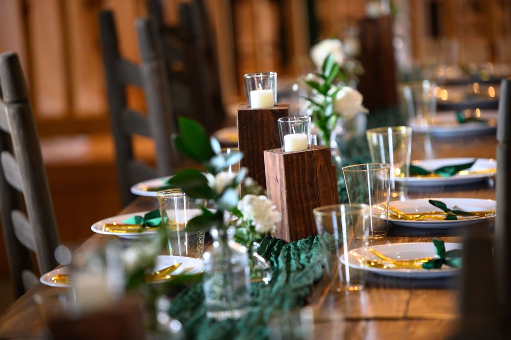 Elegant table settings with greenery centerpieces and white candles in a rustic wooden venue interior.