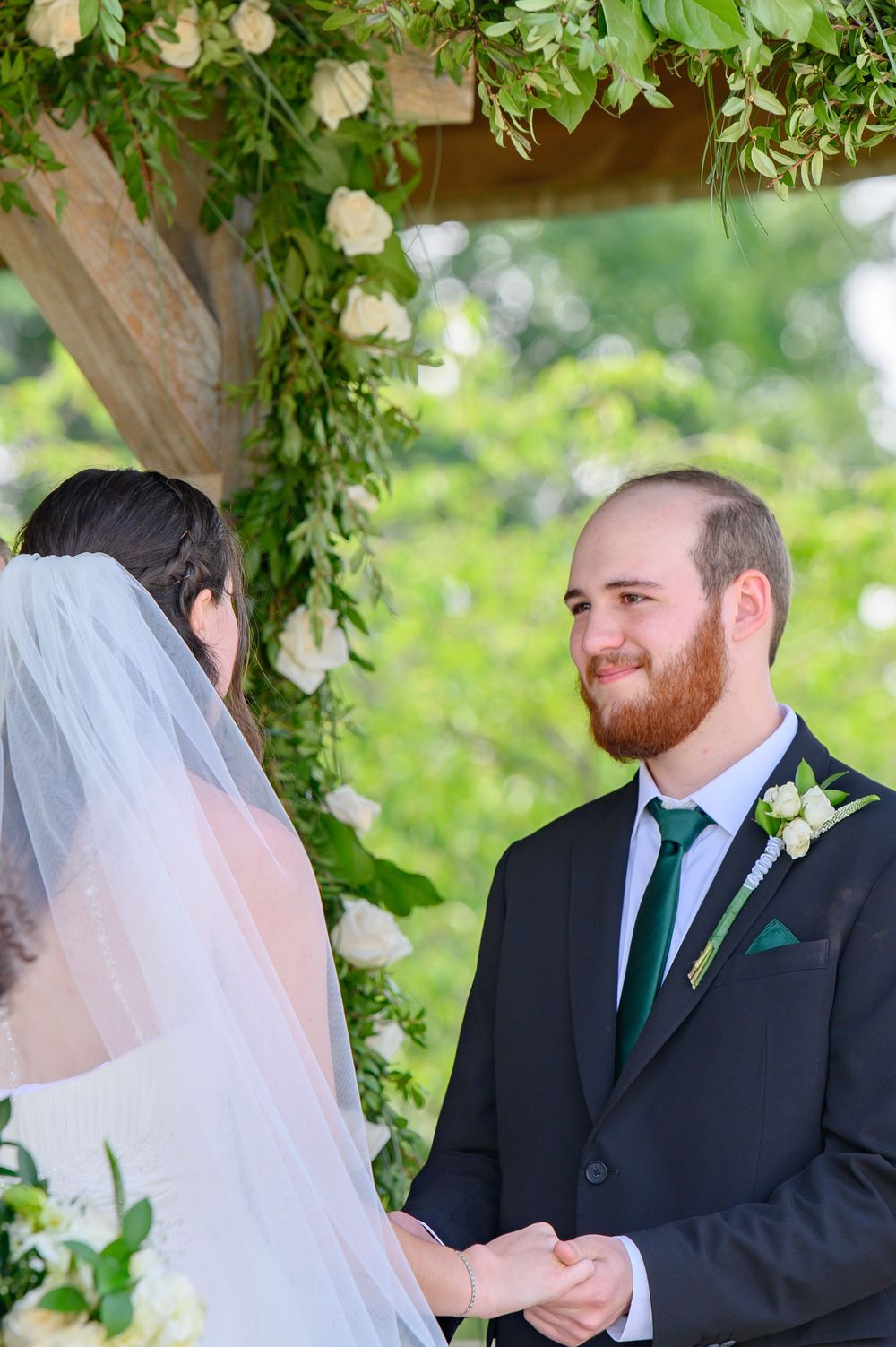 Wedding ceremony moments under green floral arch with black-suited groom and white-veiled bride.