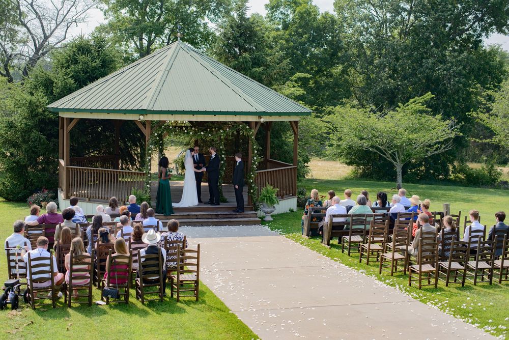 A panoramic view of an outdoor wedding venue featuring wooden chairs set up in front of a gazebo on a grassy landscape.