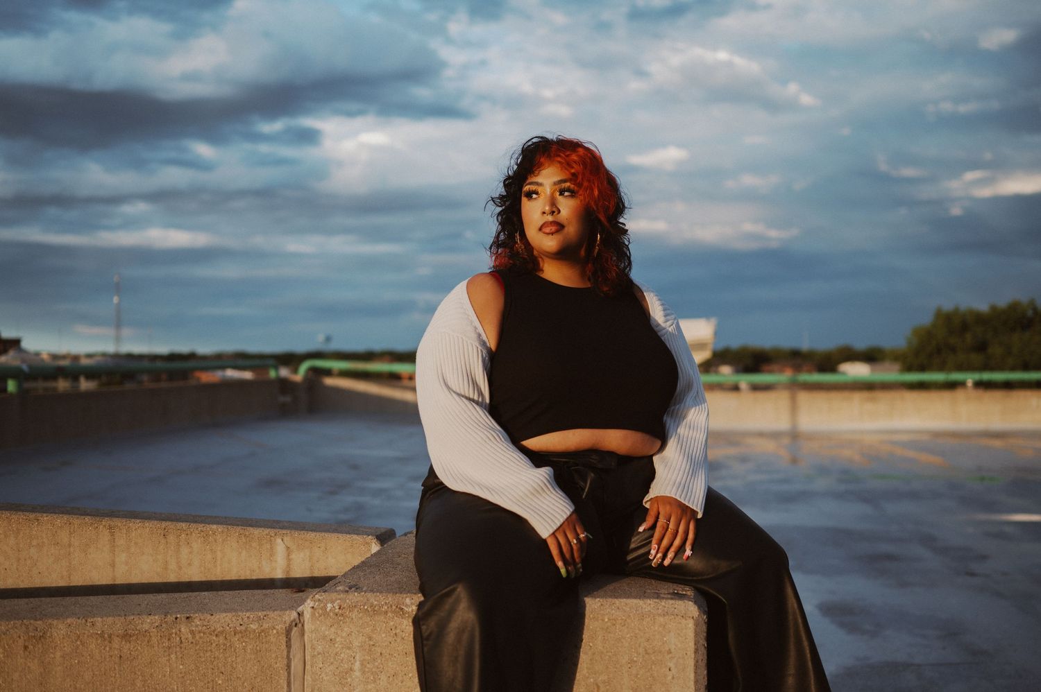 A person in casual attire sits on a concrete barrier during golden hour with a dramatic cloudy sky in the background.