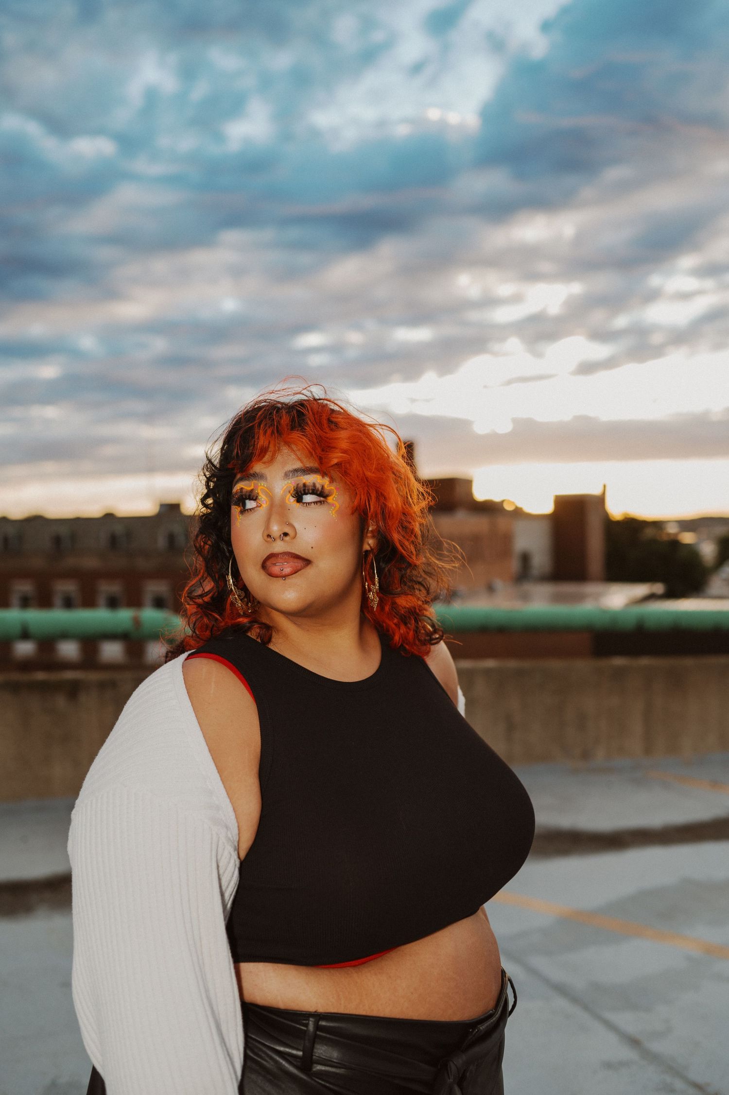 A curly-haired figure in a black crop top and white cardigan poses on a rooftop at sunset with dramatic clouds behind.