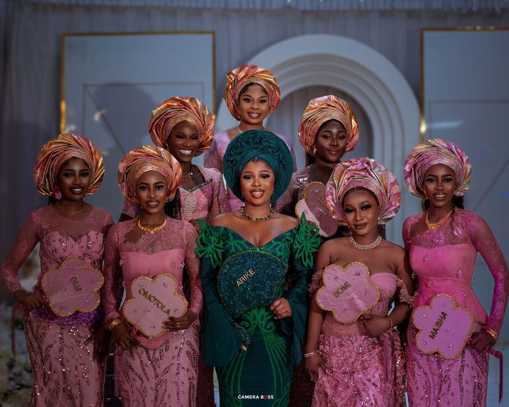 A group in traditional Nigerian attire with gele headwraps poses together wearing elegant pink and green formal dresses.