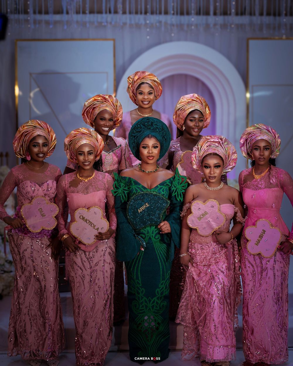A group of women in traditional Nigerian Aso Ebi attire featuring pink and emerald green gowns with matching gele headwraps at an event.