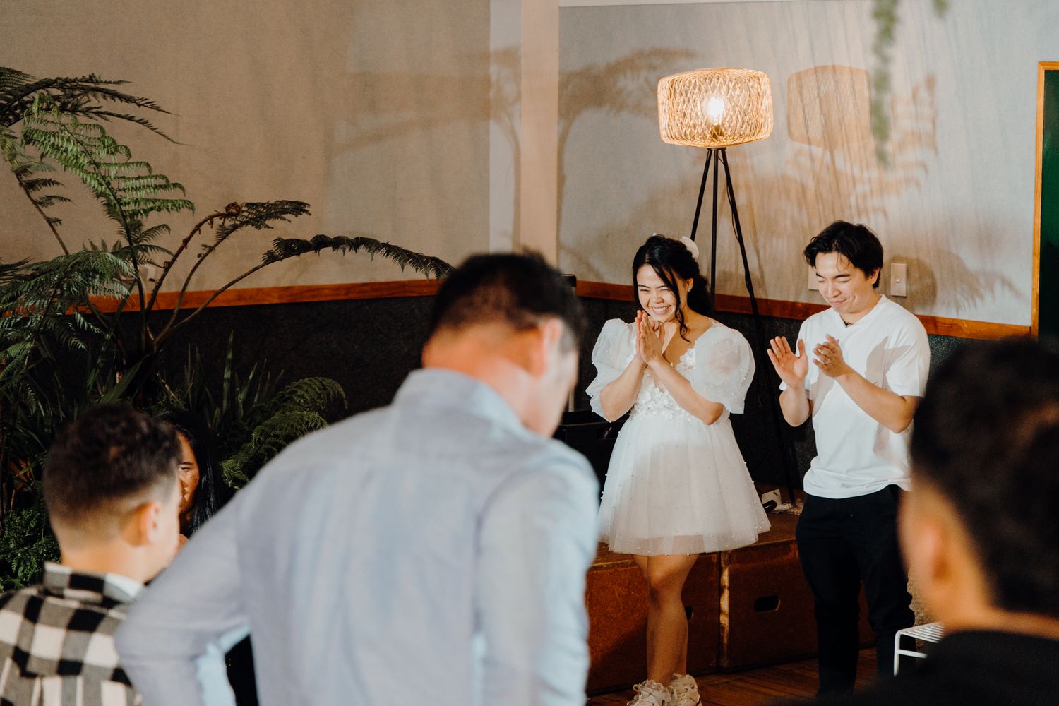 Couple performs in front of small audience in modern indoor setting with plants and lighting.
