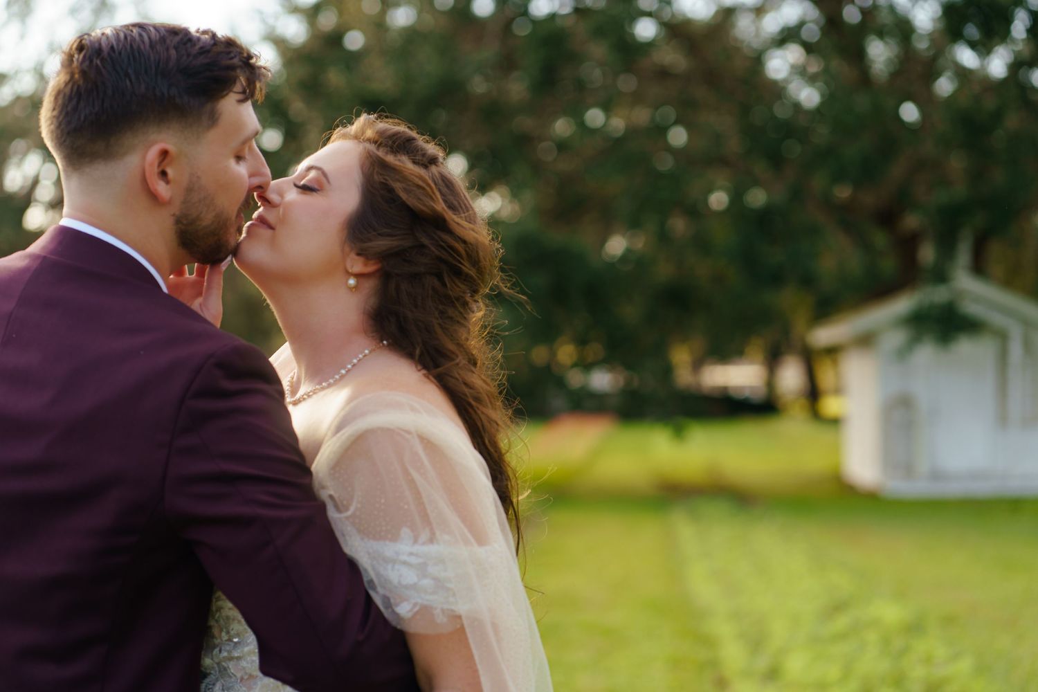 Romantic newlyweds share a tender kiss outdoors in a garden setting with trees and greenery in the background.