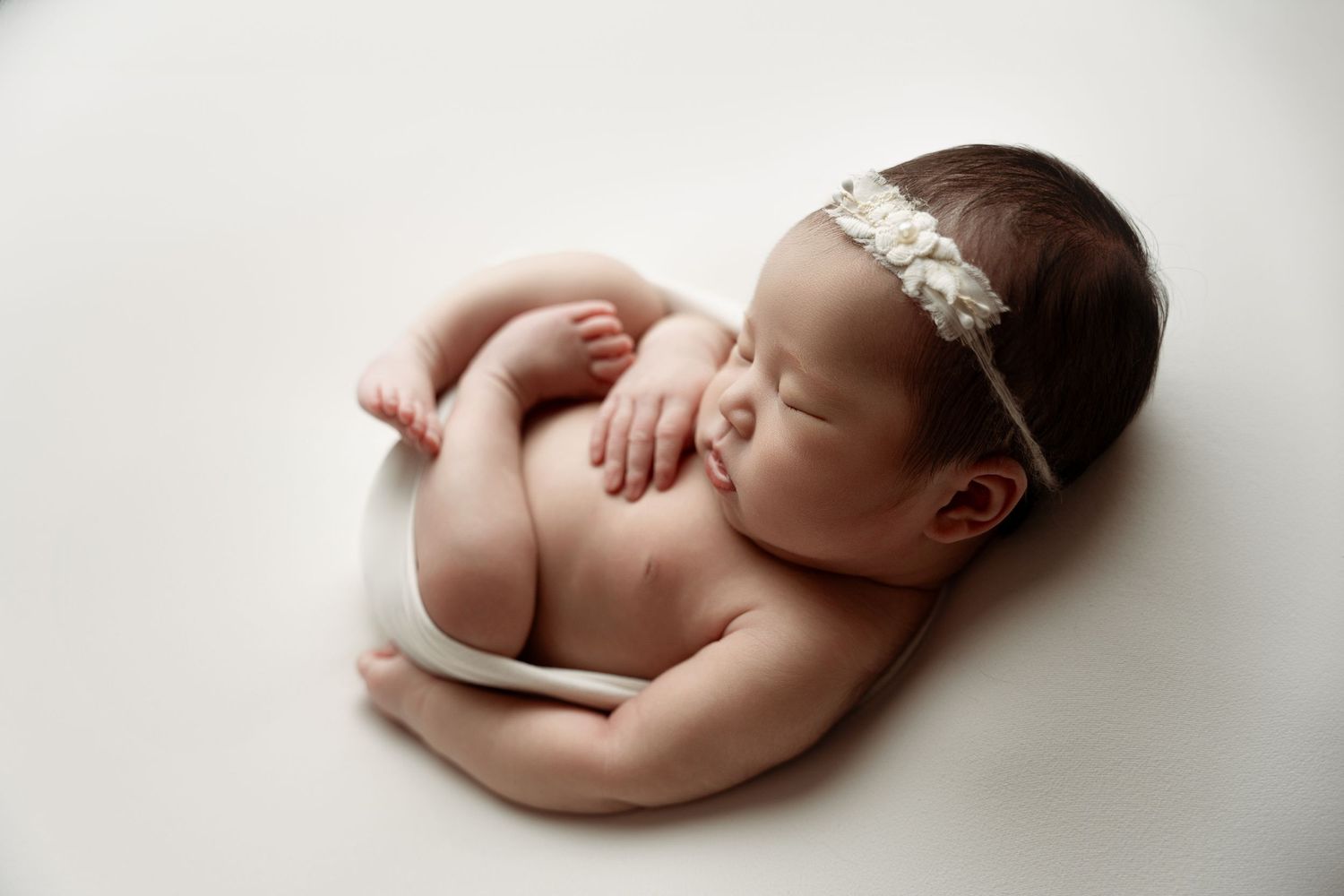 A sleeping newborn baby wearing a delicate lace headband curls up peacefully on a white backdrop during a photography session.
