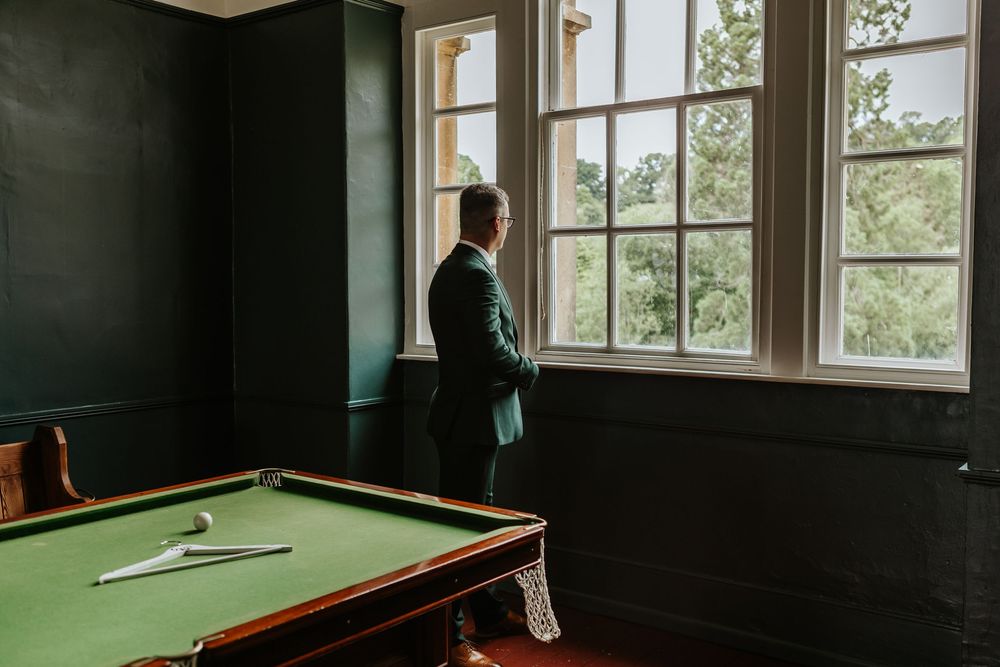 A darkly lit room with multiple windows features an empty pool table with green felt in the foreground.
