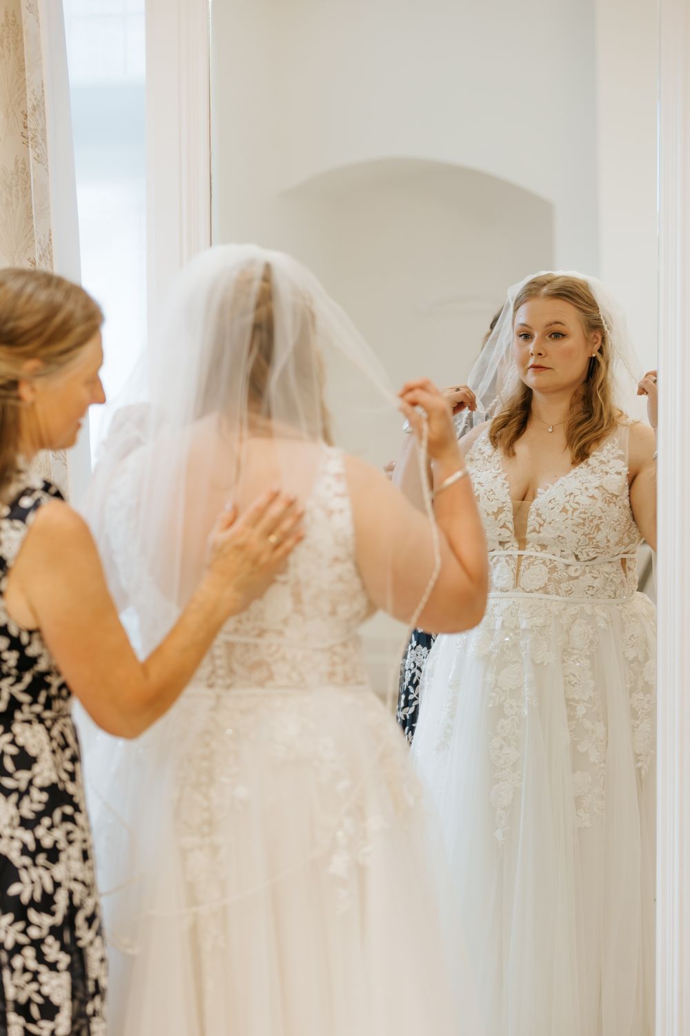 A bride tries on a lace wedding dress while attendants assist with the veil in a bridal boutique.