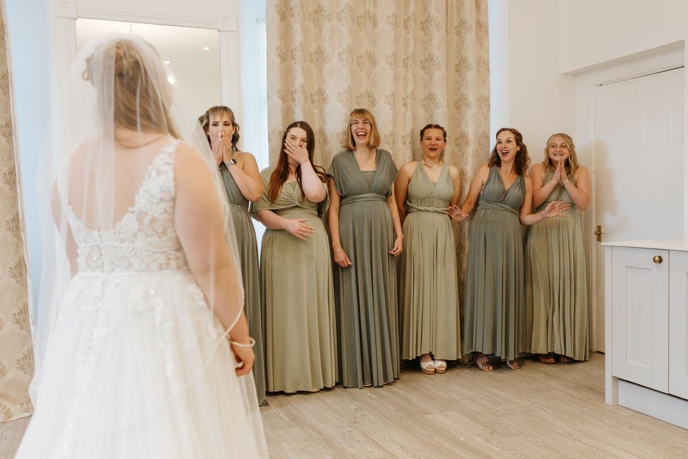 Bridesmaids in sage green dresses stand in a row beside a bride in white during wedding preparations.