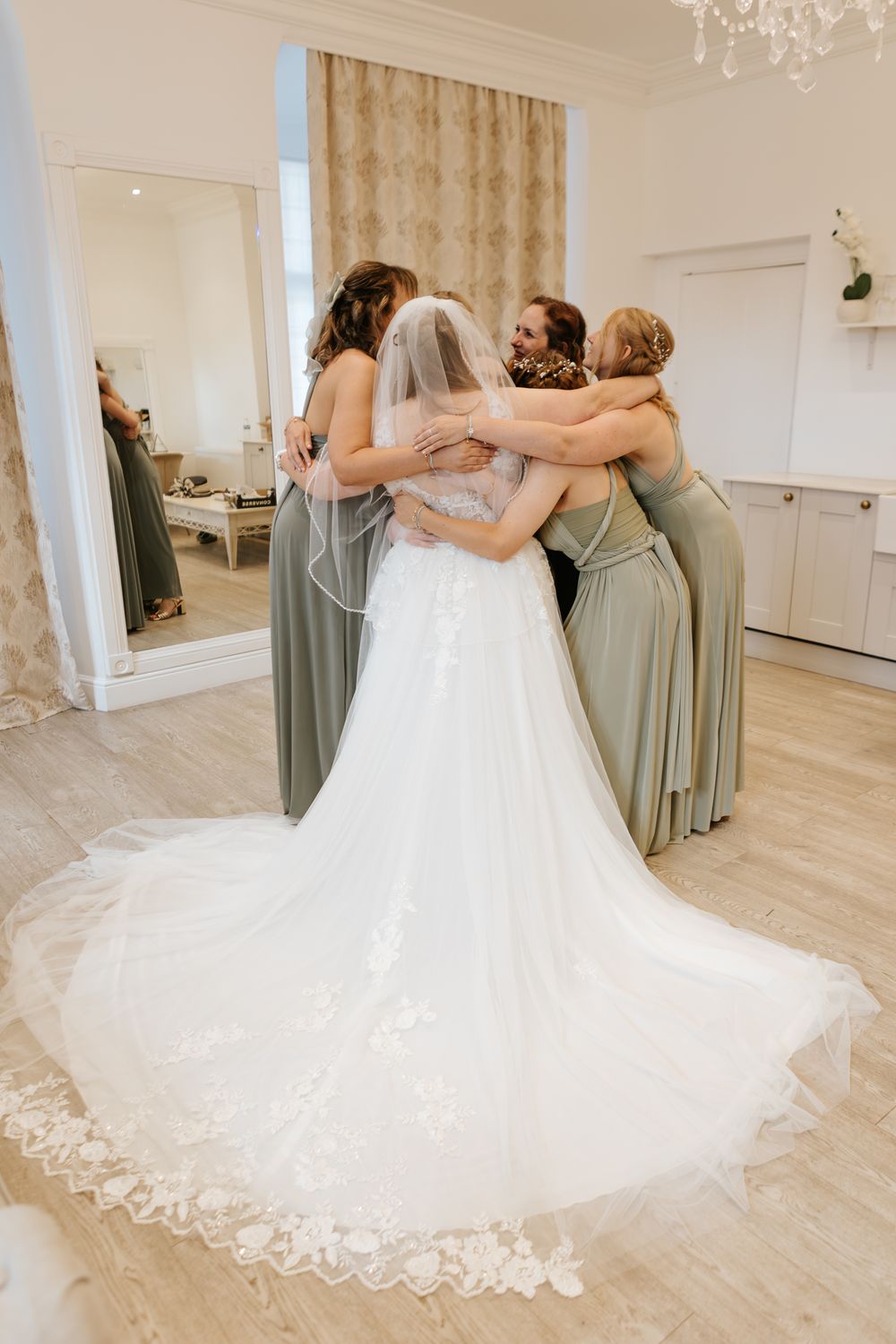 A group in sage green bridesmaid dresses embraces in a circle while a white wedding gown train spreads out gracefully.