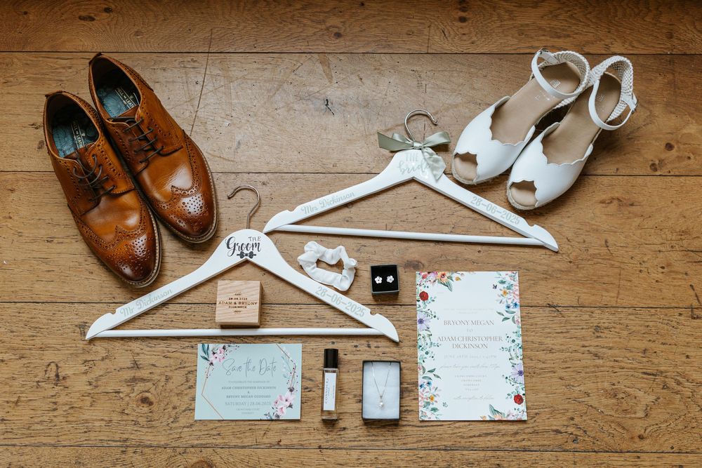 Wedding accessories including brown leather shoes, white heels, hangers, and stationery laid out on wooden flooring.