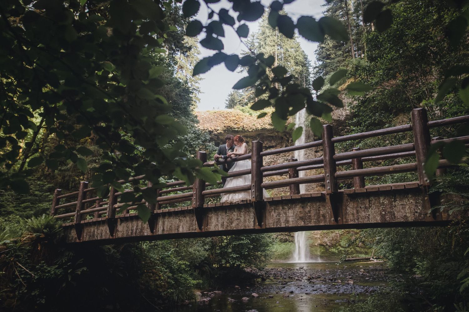 A rustic wooden footbridge spans over a creek in a lush green forest setting while a couple enjoys the natural scenery.