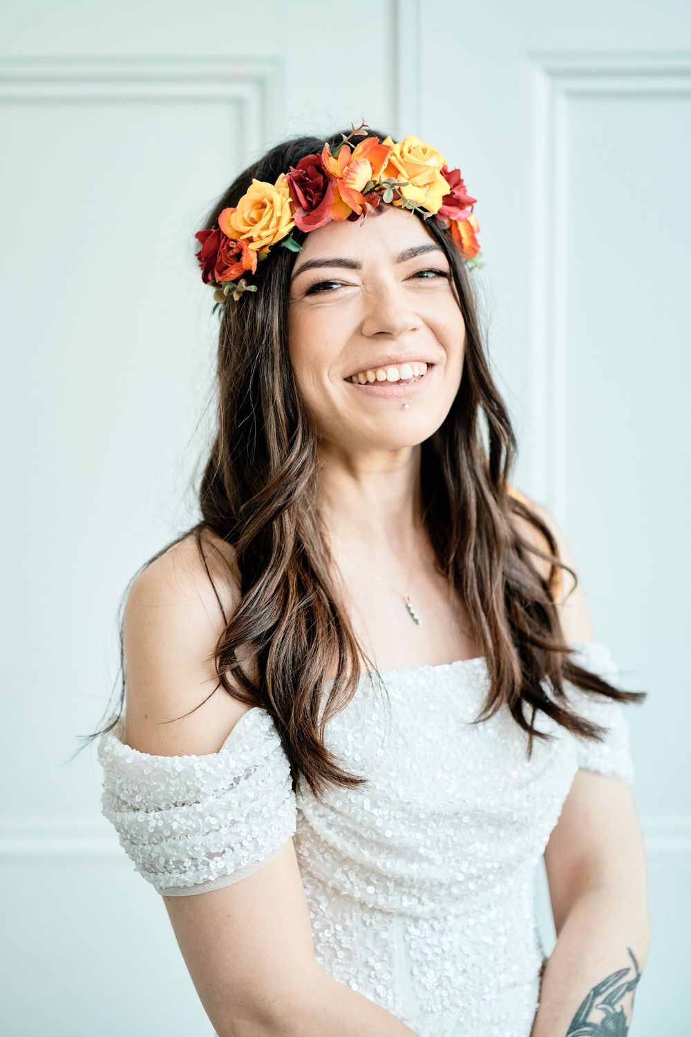 Portrait of someone wearing a white lace dress and colorful floral crown with orange and pink roses smiling against white wall.
