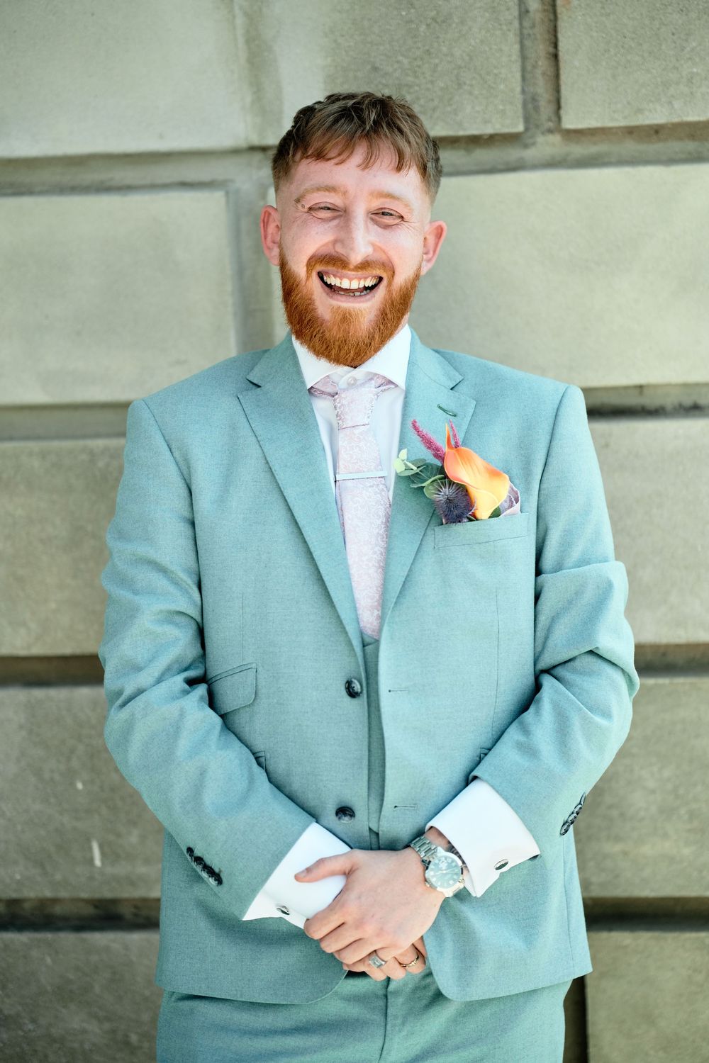 Groom wearing a mint green suit and floral boutonniere stands smiling in front of stone steps.