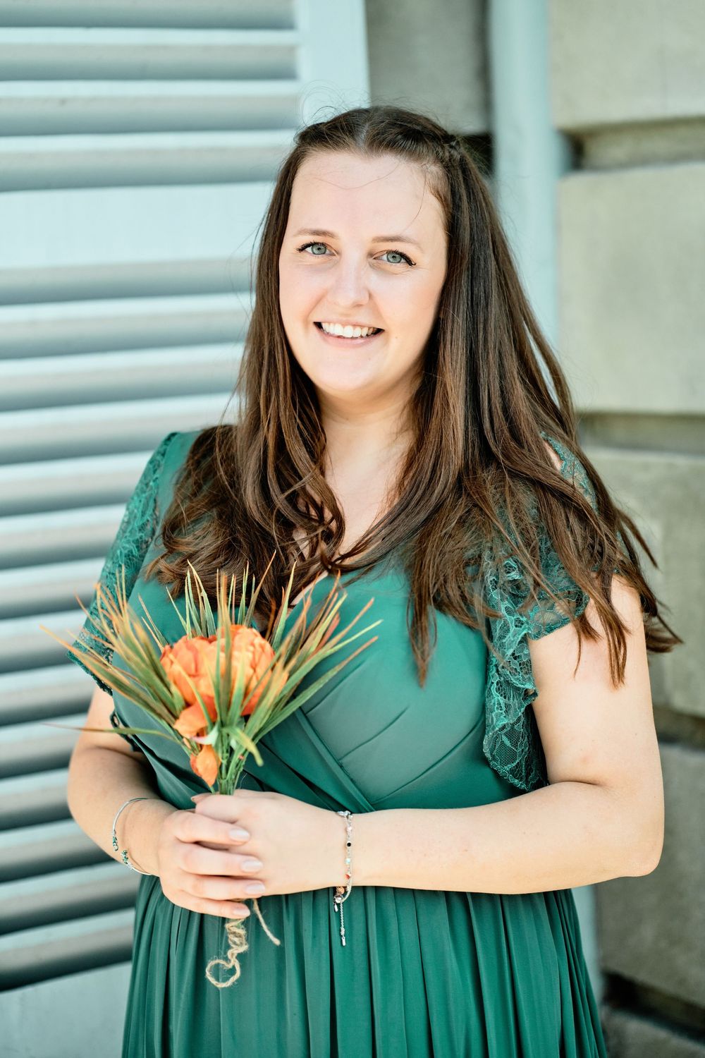 Person in an emerald green dress holds a small bouquet while smiling outdoors near shutters.