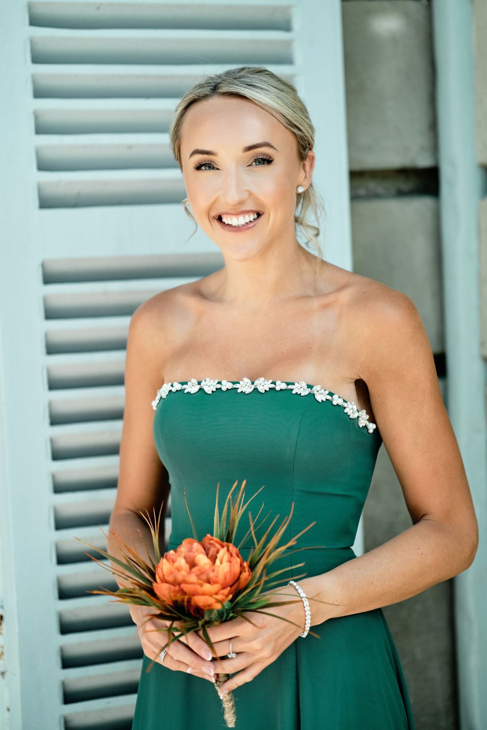 Woman in elegant emerald green strapless dress with floral trim holds a coral peony bouquet against blue shutters.