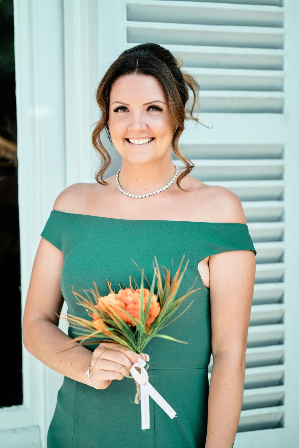 Person wearing an emerald green off-shoulder dress holding orange tropical flowers against blue shutters.