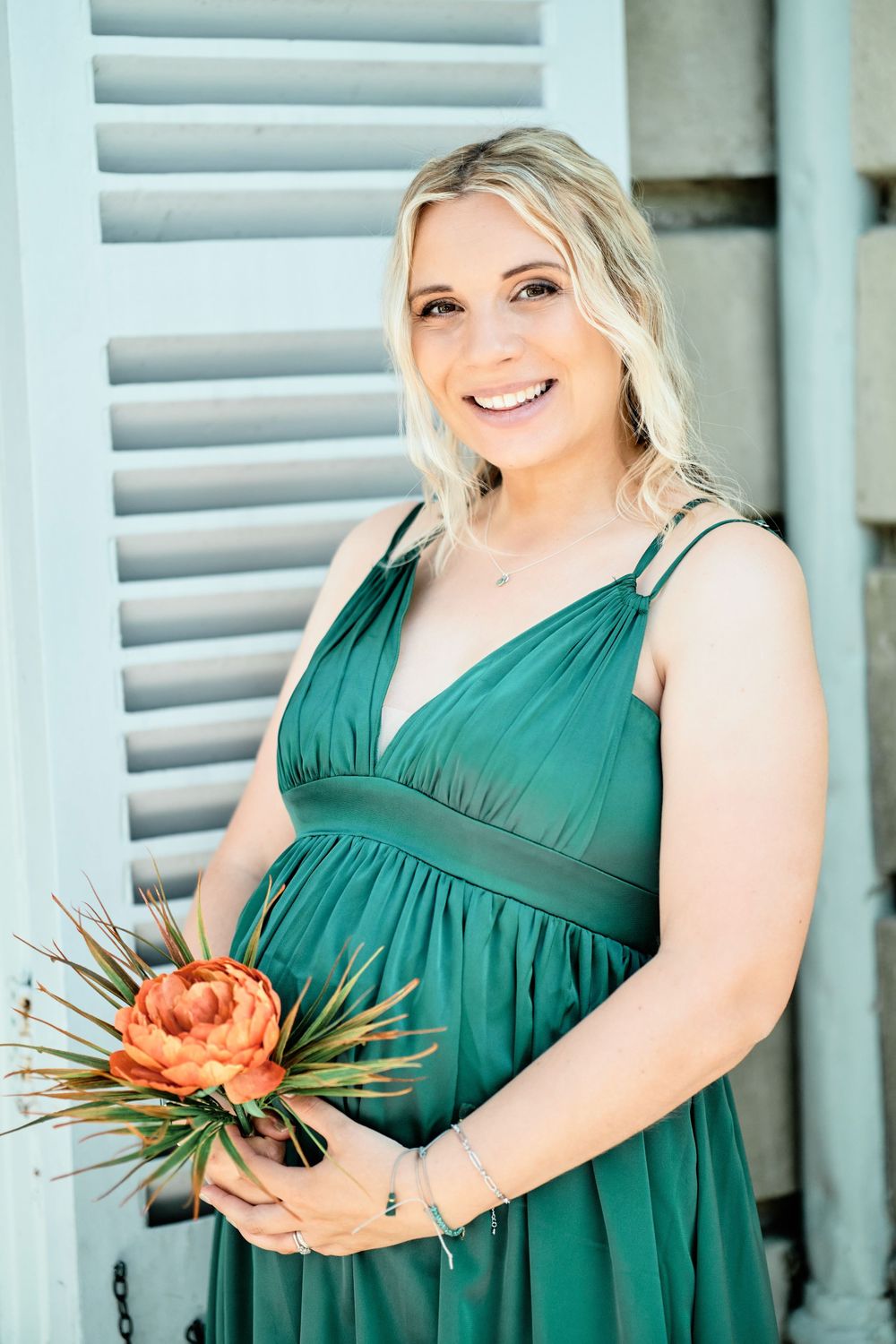 Expectant woman in emerald green maternity dress holds orange protea flower against blue shuttered background.