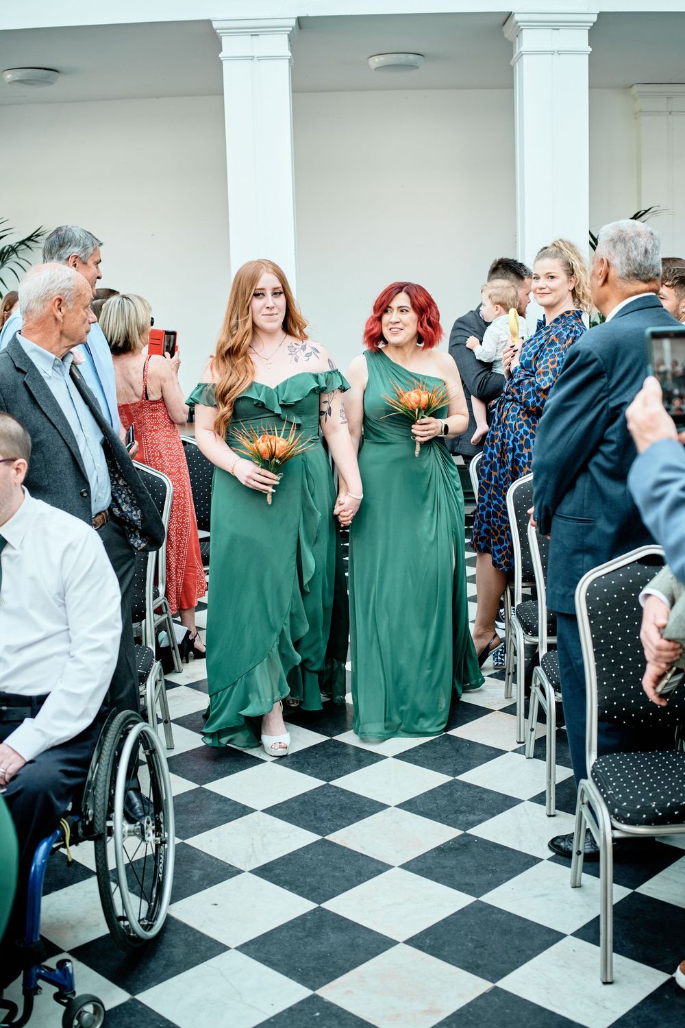 Wedding party guests in green dresses and formal wear mingle on a black and white checkered floor.